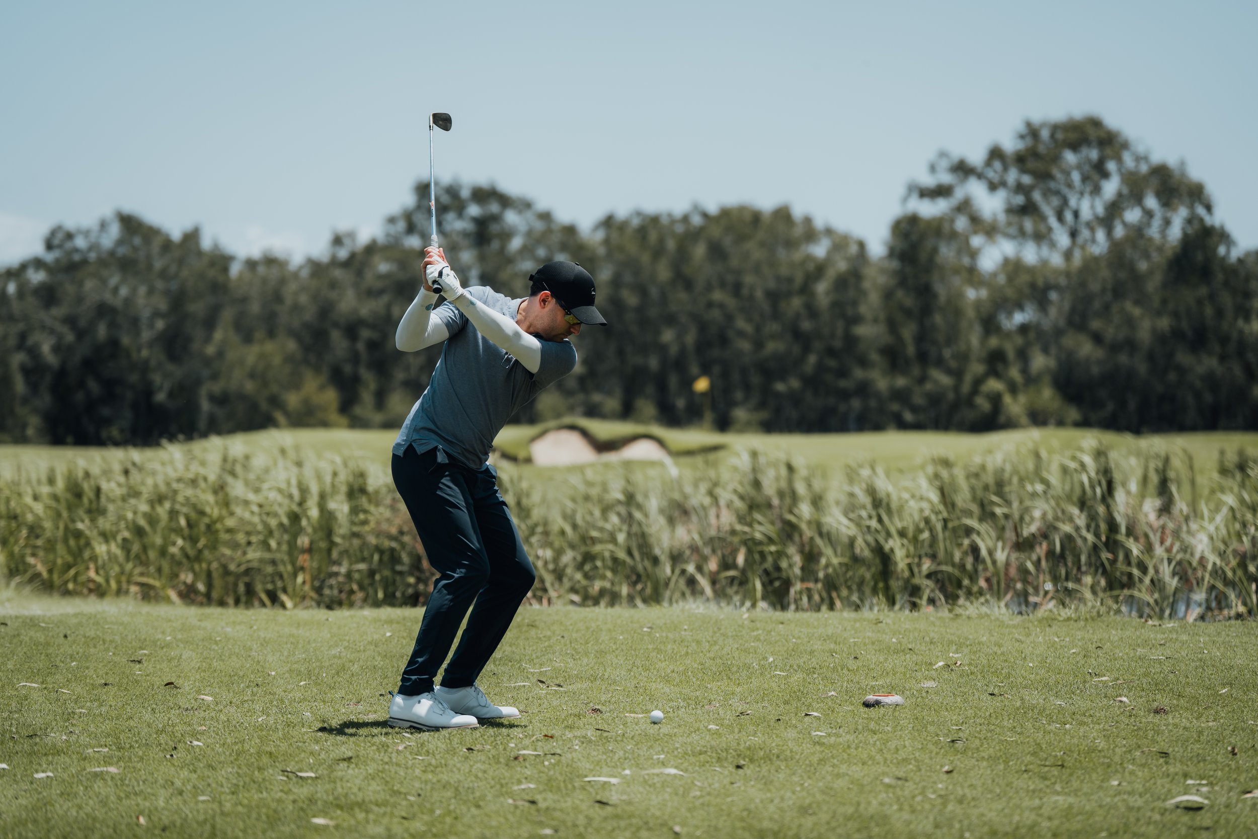 A man in a hat and sunglasses takes a golf swing on a lush golf course with trees and a pond in the background.