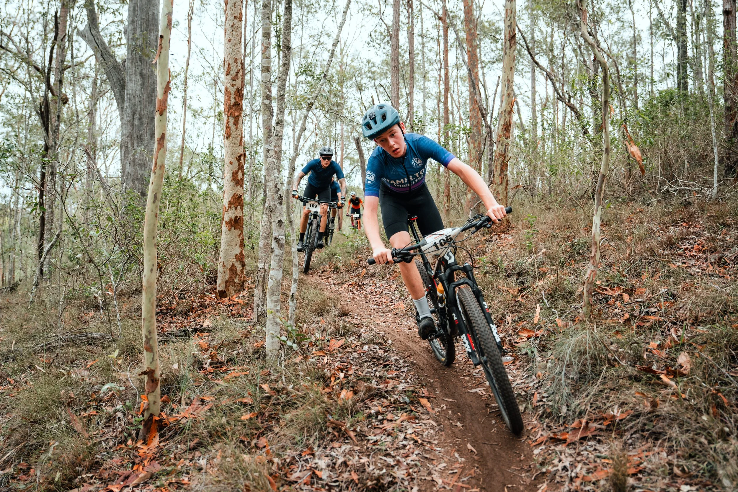 Group of mountain bikers riding on a dirt trail through a wooded forest.