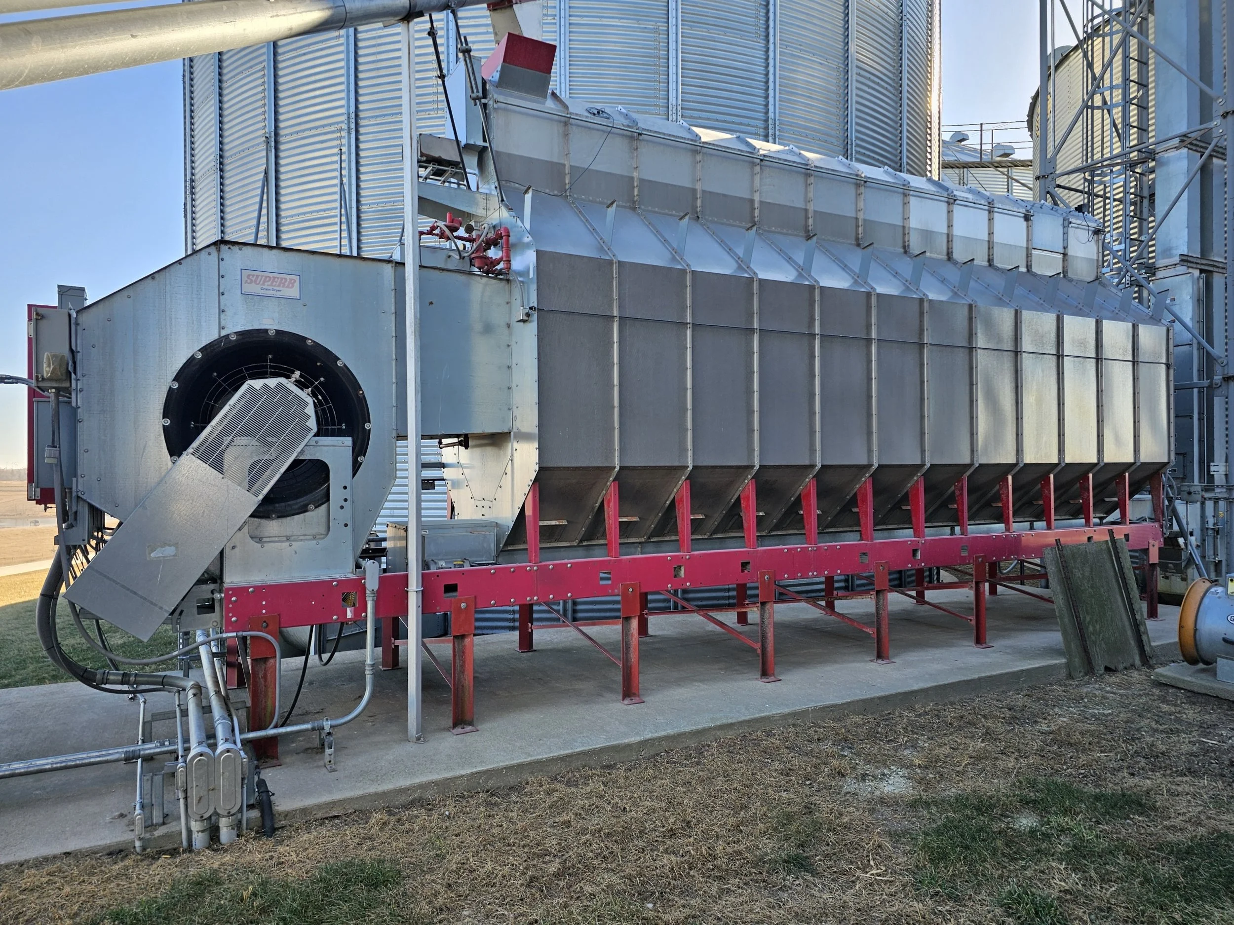 Industrial equipment with metal ducts, pipes, and a fan system, situated on a concrete pad, with grain silos in the background.