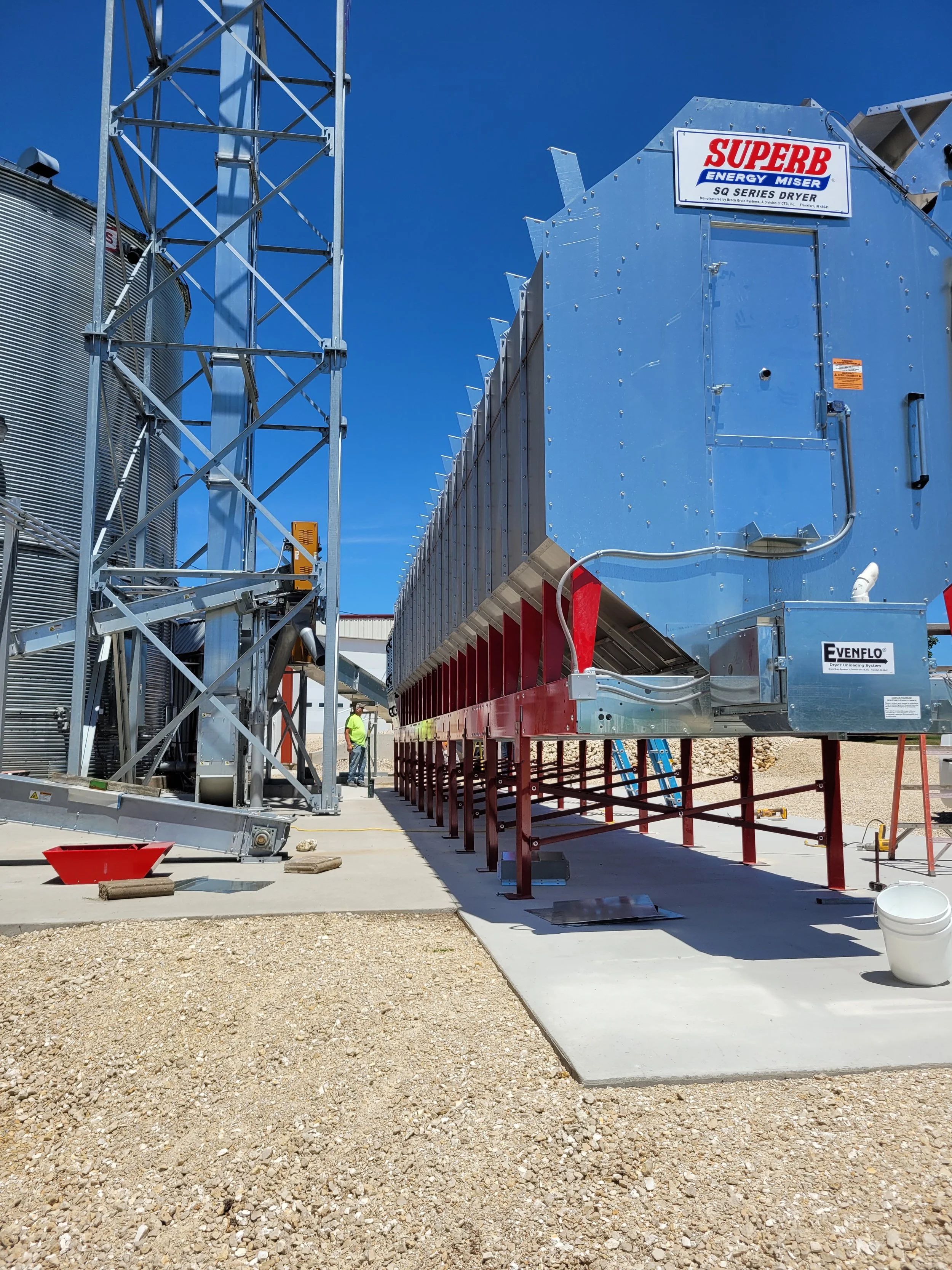 Large industrial machine labeled 'Superb Energy Miser SQ Series Dryer' installed outdoors on concrete, with a person in a high-visibility vest nearby and construction materials around.