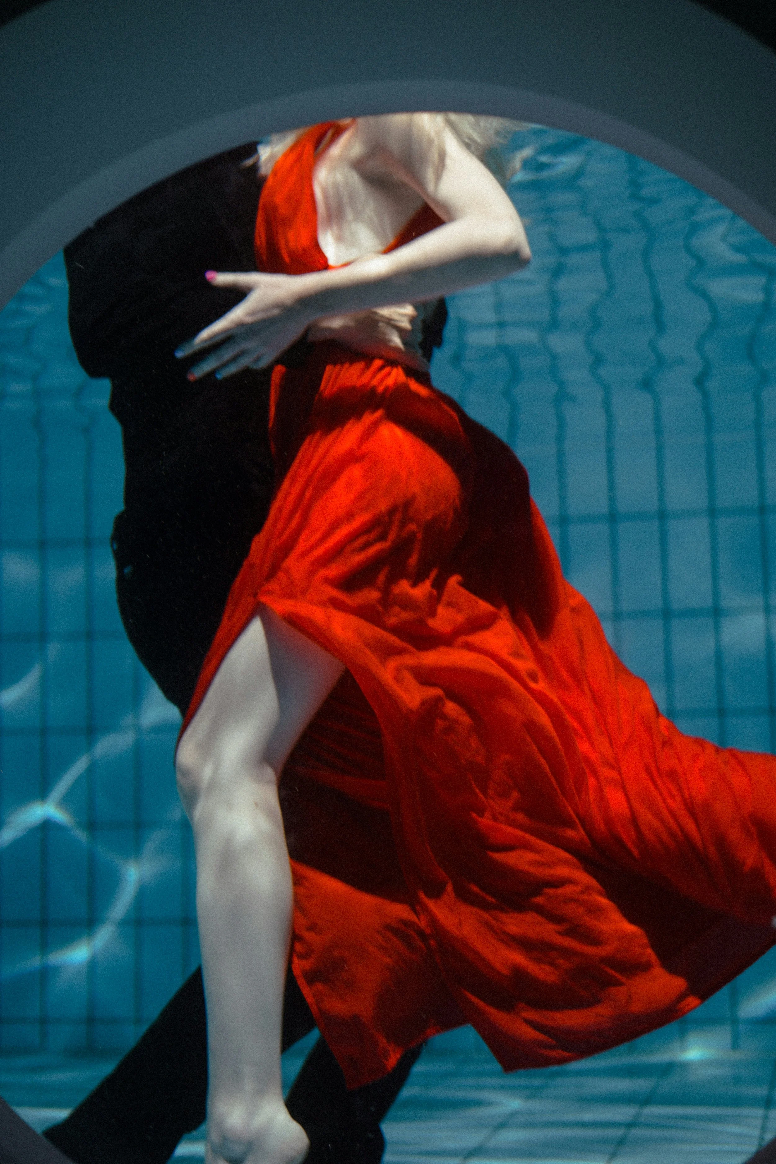 A person in a red dress and black outfit underwater, viewed through a porthole.