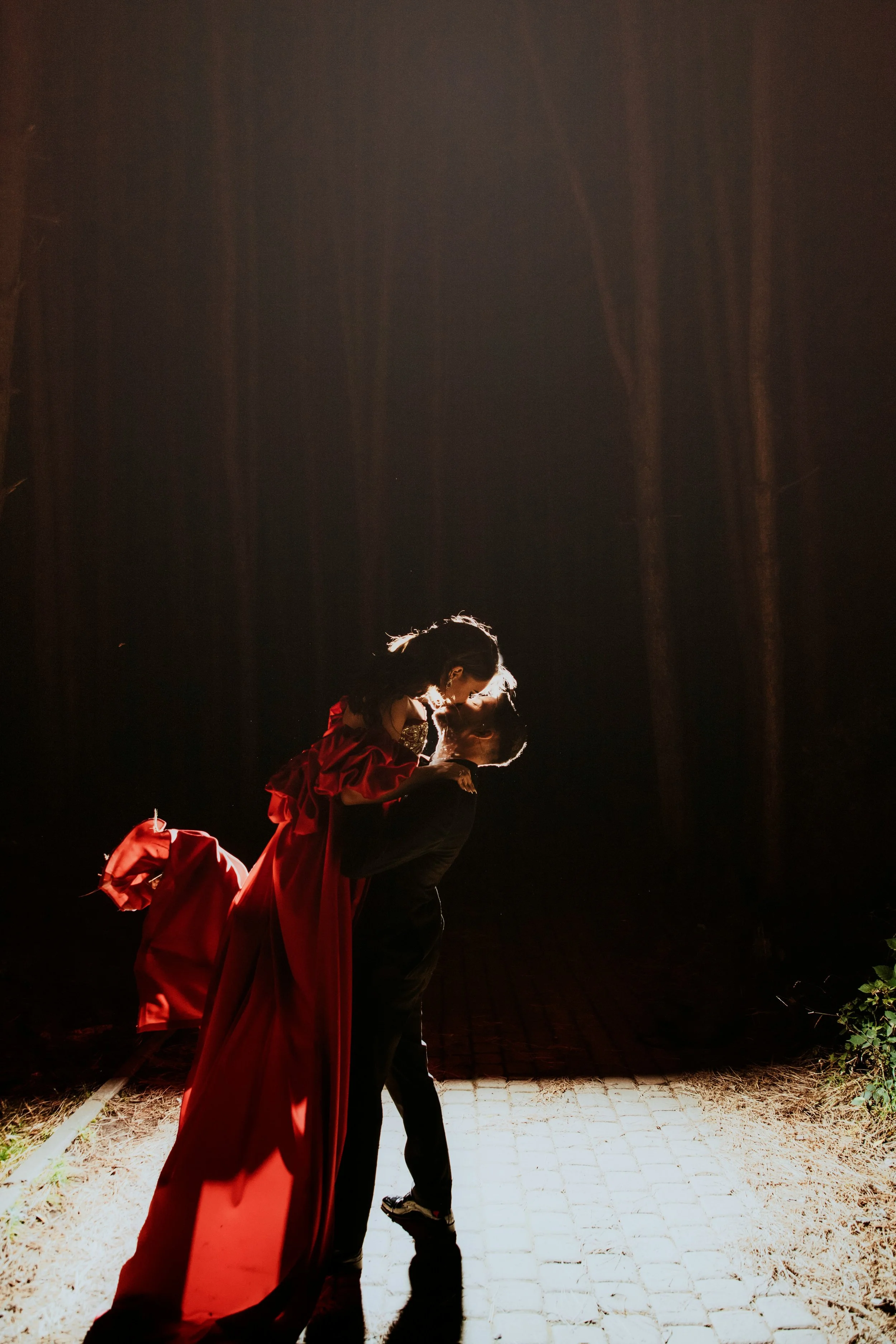 A couple sharing a kiss at night, with backlit illumination highlighting their silhouettes. The woman is wearing a long, flowing red dress, and the man is dressed in dark clothing. They are standing on a paved surface with a dark forest background.