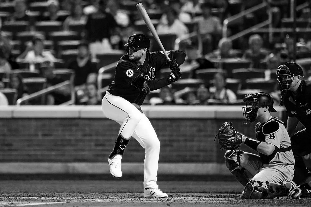 A black and white photo of a baseball game showing a player at bat, a catcher in a squatting position, and an umpire behind home plate, with spectators in the background.