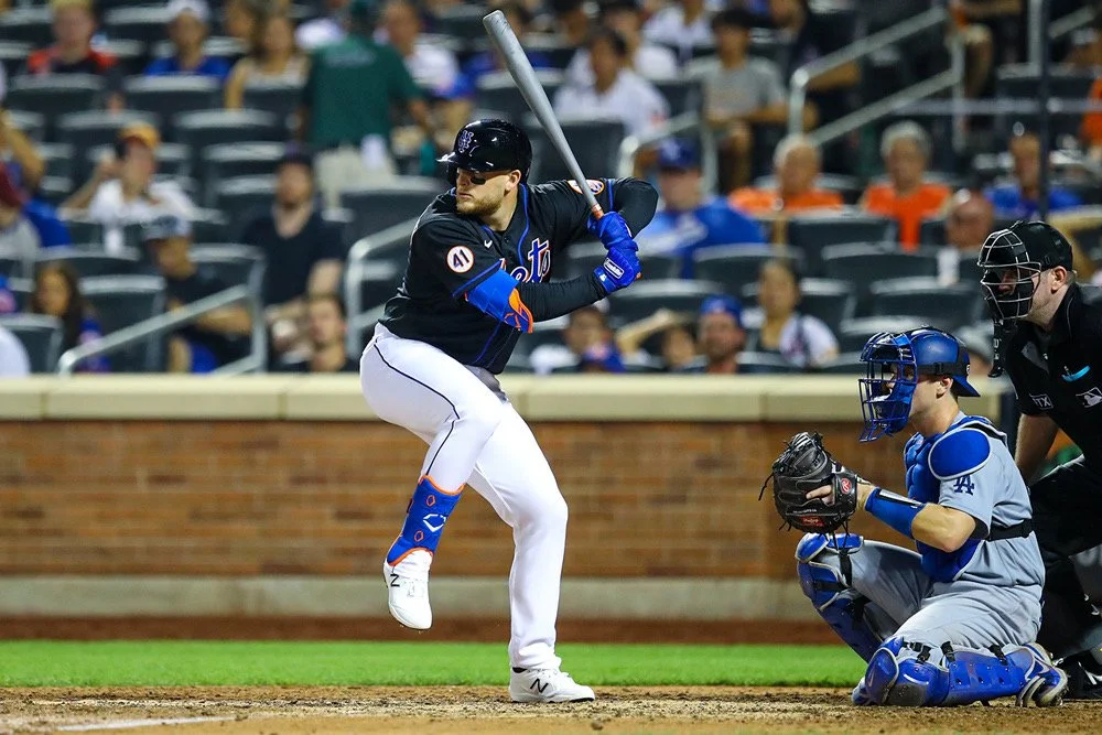 A baseball player from the New York Mets batting, with a catcher and umpire behind him, and spectators in the background.