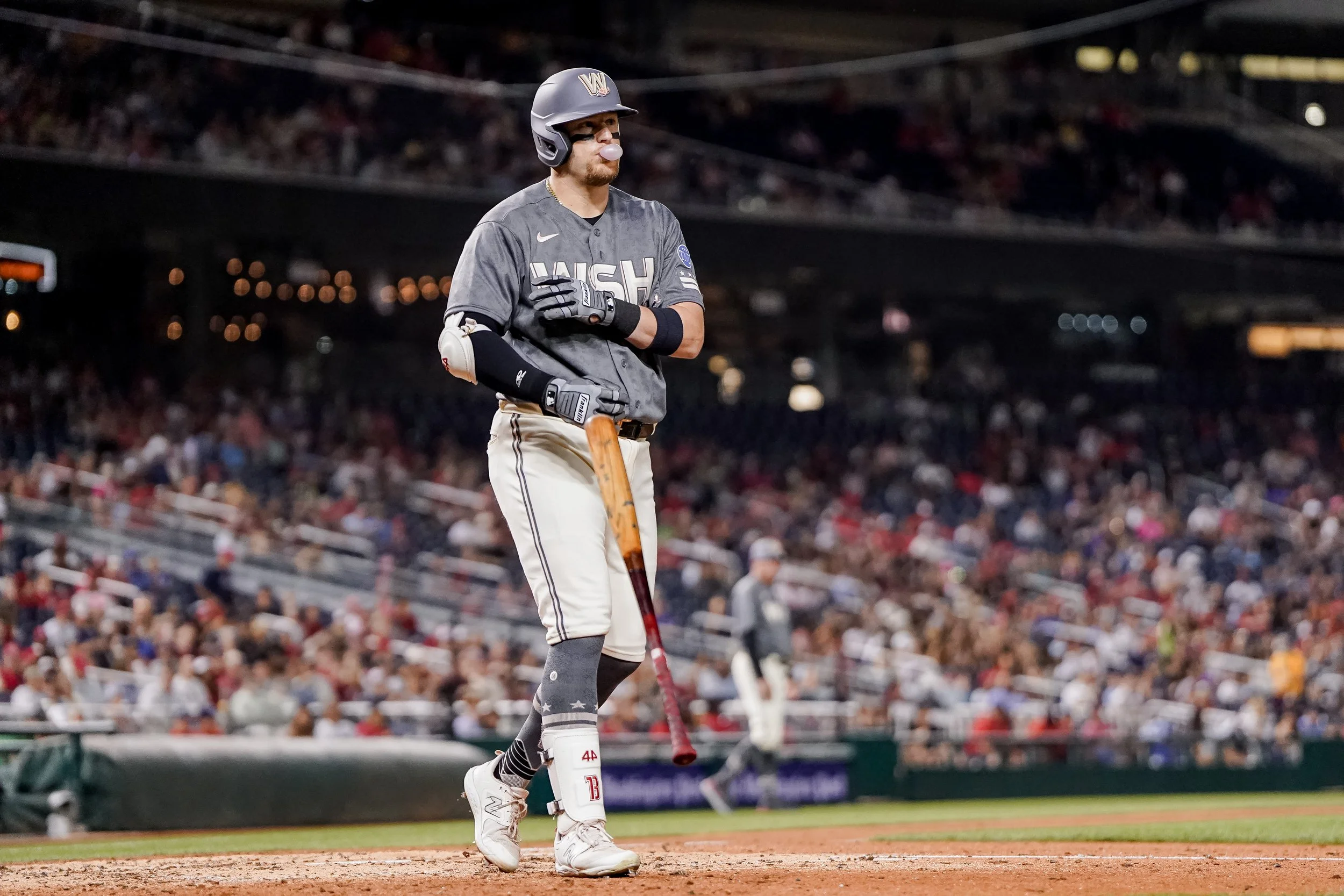A baseball player wearing a dark gray uniform and helmet standing on home plate with a bat in hand during a game, with a crowd in the stands in the background.