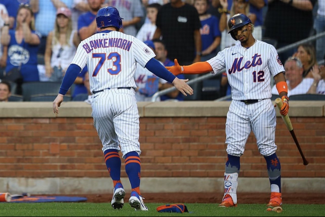 Two New York Mets baseball players, one in a blue helmet and the other in a cap, high-five on the field during a game with fans in the background.