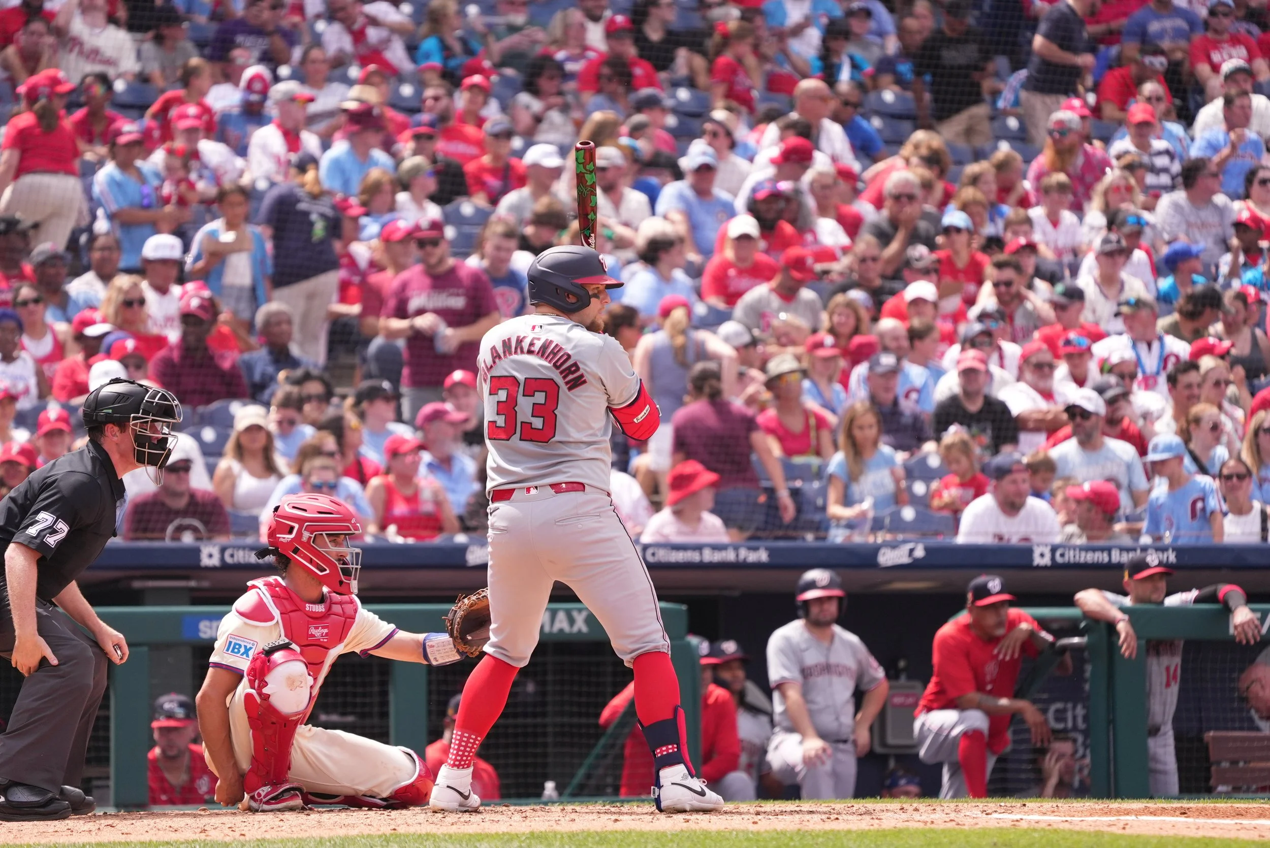 A baseball player in a gray and red uniform with the number 33 and the name 'Flankenhorn' at bat, a catcher in a white uniform crouching behind home plate, and an umpire in black crouching, with a crowd of fans wearing red, white, and blue in the background.