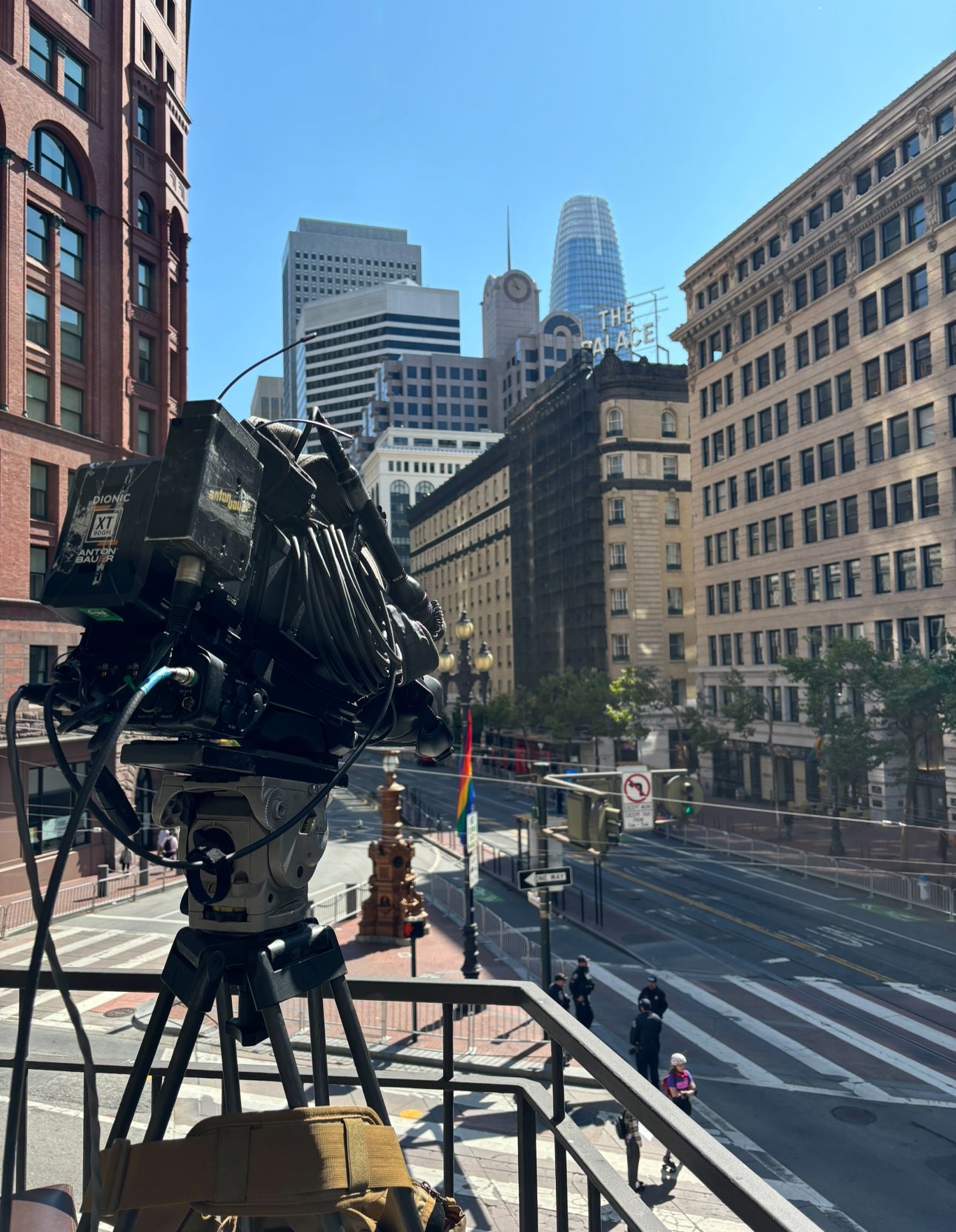 A professional camera mounted on a tripod on a balcony overlooking a city street with tall buildings, traffic lights, pedestrians, and a rainbow flag.