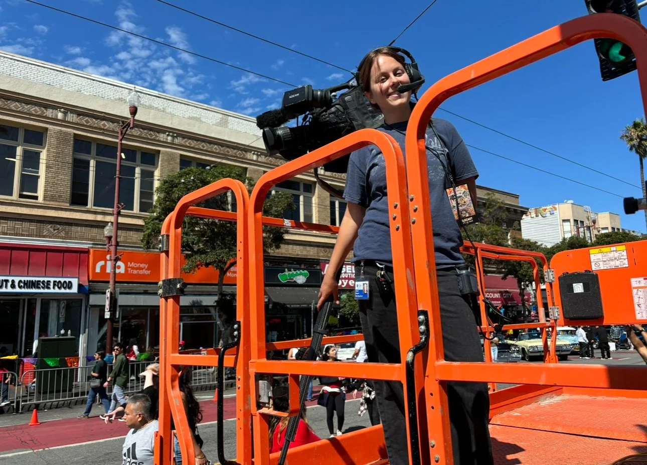 A woman operating a camera on an orange lift in a city street with shops, pedestrians, and a clear blue sky.