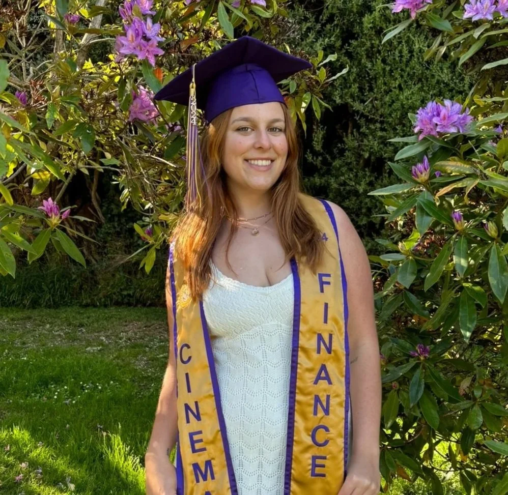 A young woman in a graduation cap and gown, smiling outdoors among purple flowering bushes.