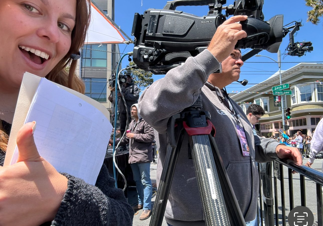 A woman smiling and holding a notebook while filming at an outdoor event with a professional camera operator beside her. The scene includes other people, street signs, and a clear blue sky.