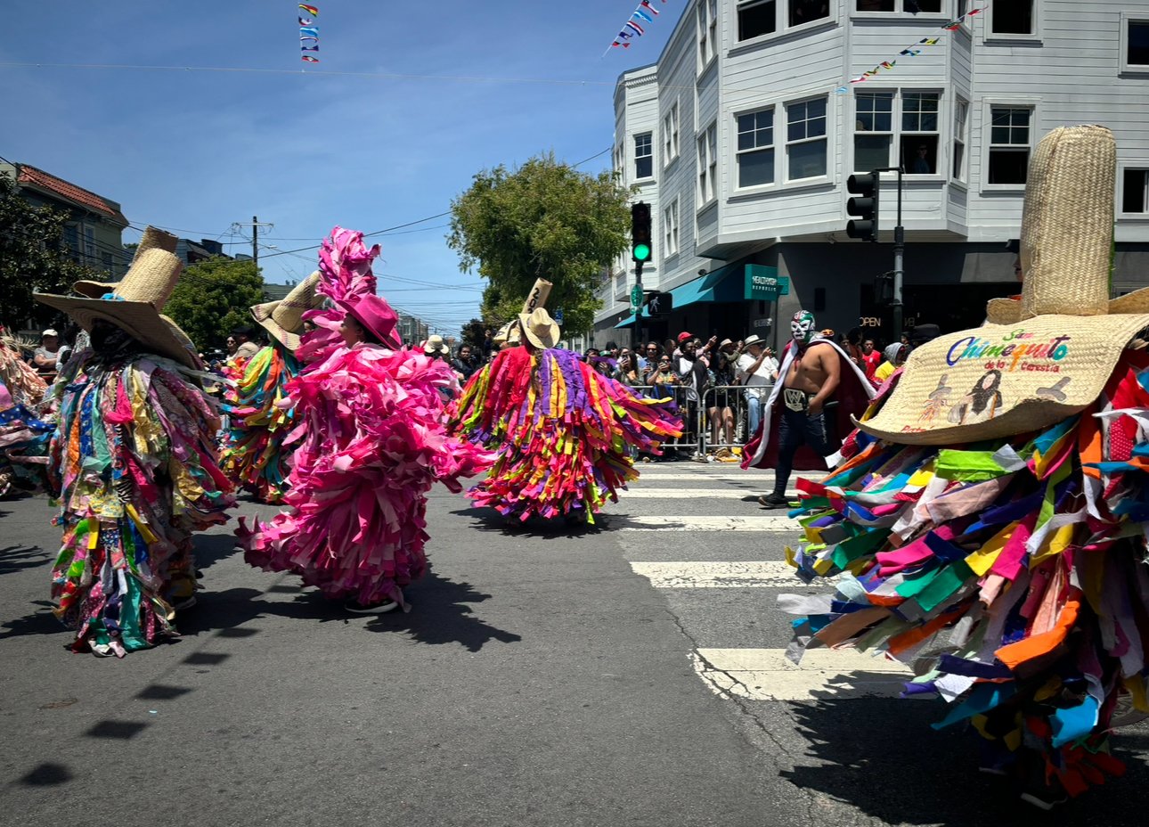 People in colorful costumes celebrating in a parade on a city street, with onlookers behind barriers and buildings in the background.