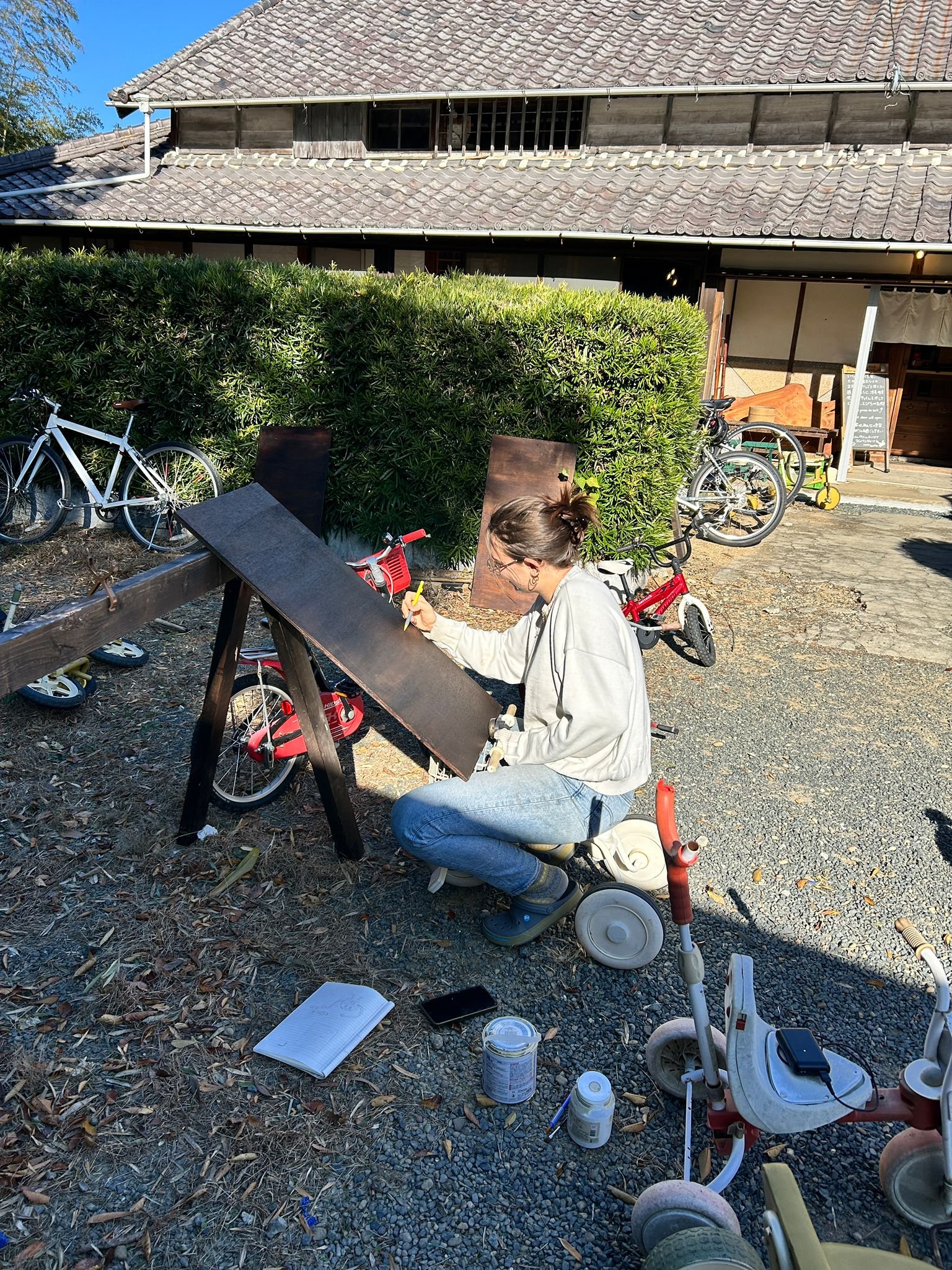 Une femme peint sur une toile en plein air, entourée de vélos, avec une maison traditionnelle en arrière-plan.