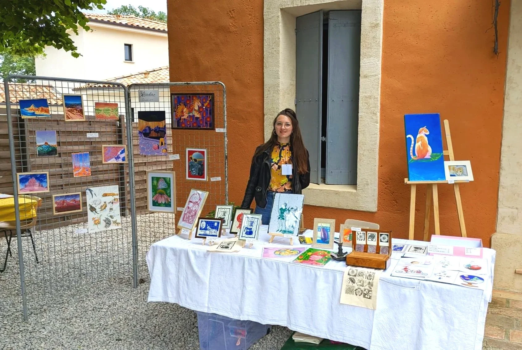 Une jeune femme (blandine guillet) à une exposition d'art en plein air, devant une table avec diverses œuvres d'art comme des peintures et des dessins, avec un panneau de présentation en arrière-plan et un mur orange avec une fenêtre.