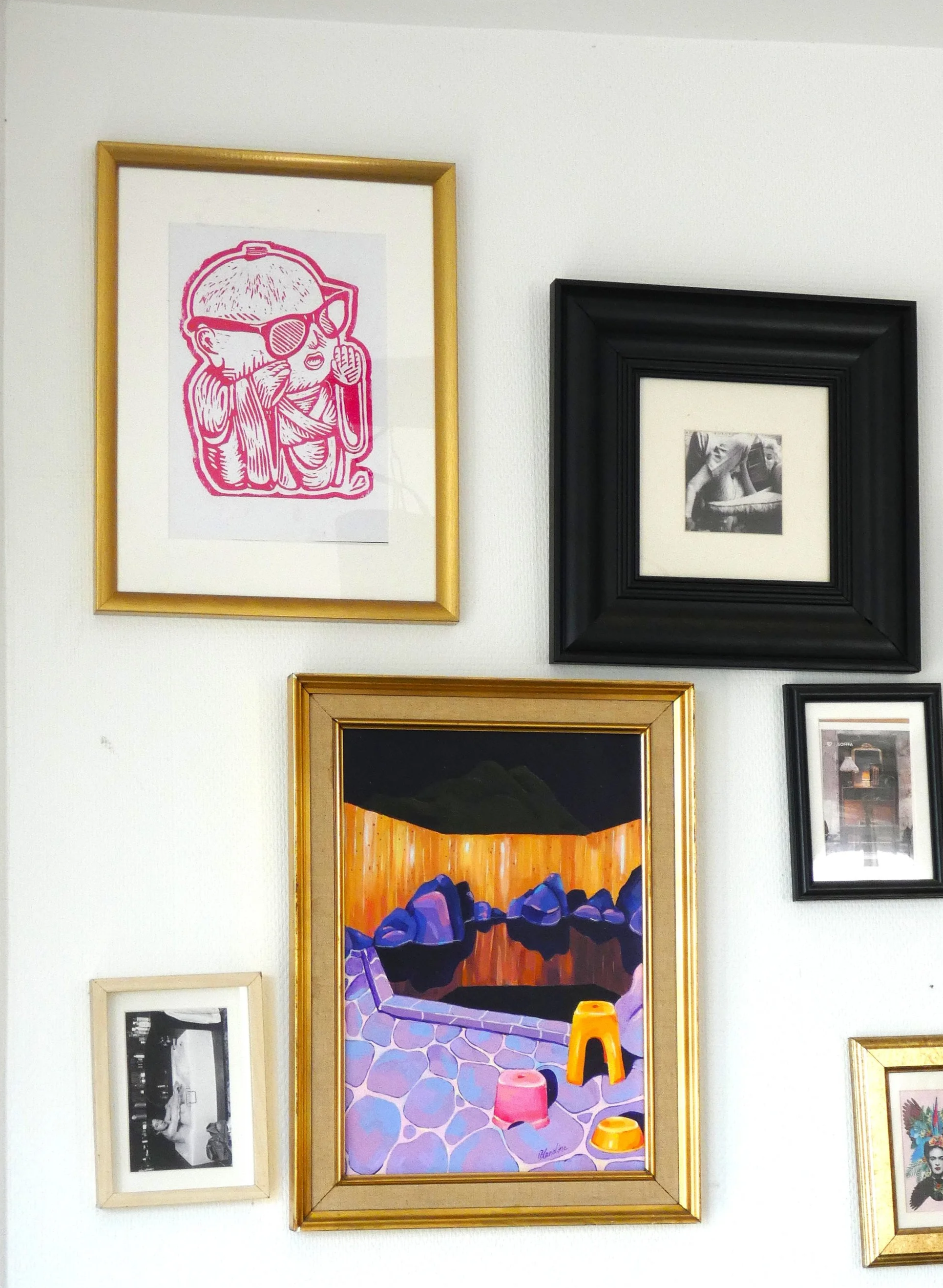 Un mur blanc orné de plusieurs cadres contenant diverses œuvres d'art, y compris une illustration rose d'un personnage portant des lunettes de soleil, une photo noir et blanc d'une femme, une peinture colorée d'un paysage avec lacs et montagnes, une petite photo en noir et blanc, et une autre image colorée en partie visible.