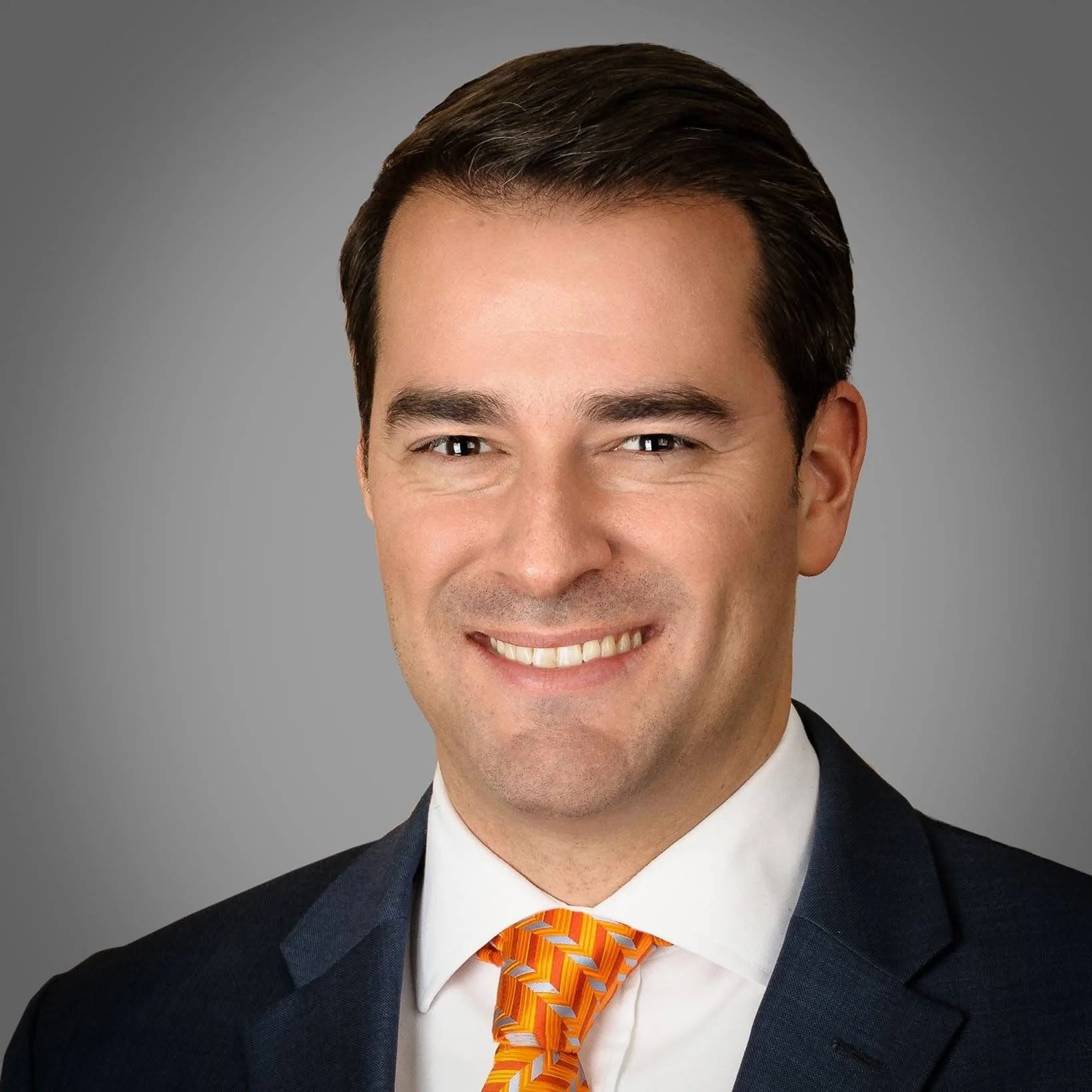 Professional headshot of a smiling man wearing a dark suit, white shirt, and an orange patterned tie against a gray background.