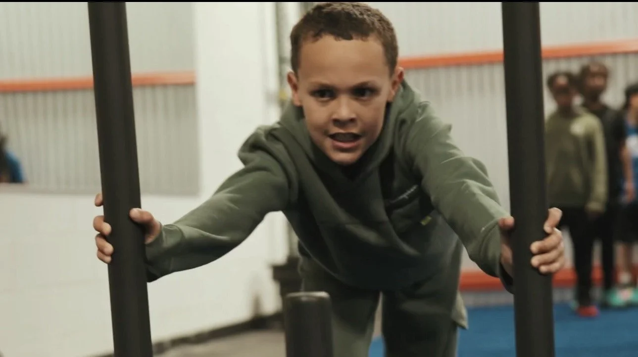 Young boy pushing a metal pole during a physical activity or gym class, with other children in the background.