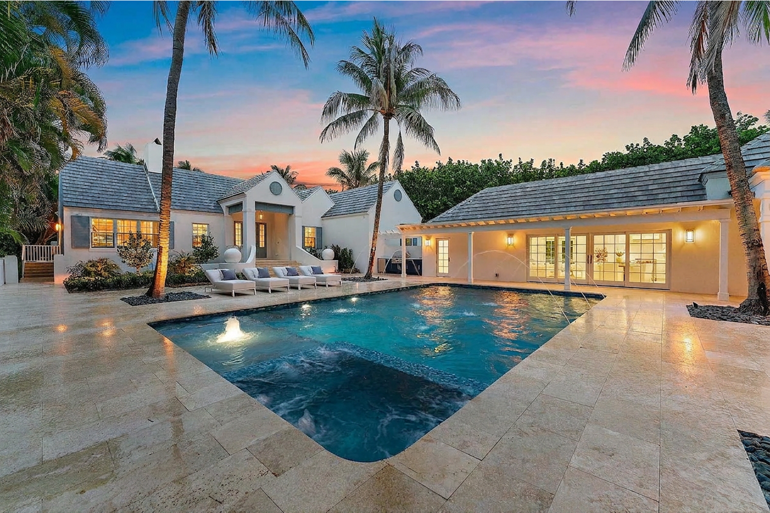 A luxurious backyard with a swimming pool, surrounded by palm trees, patio chairs, and a house with lit windows at sunset.