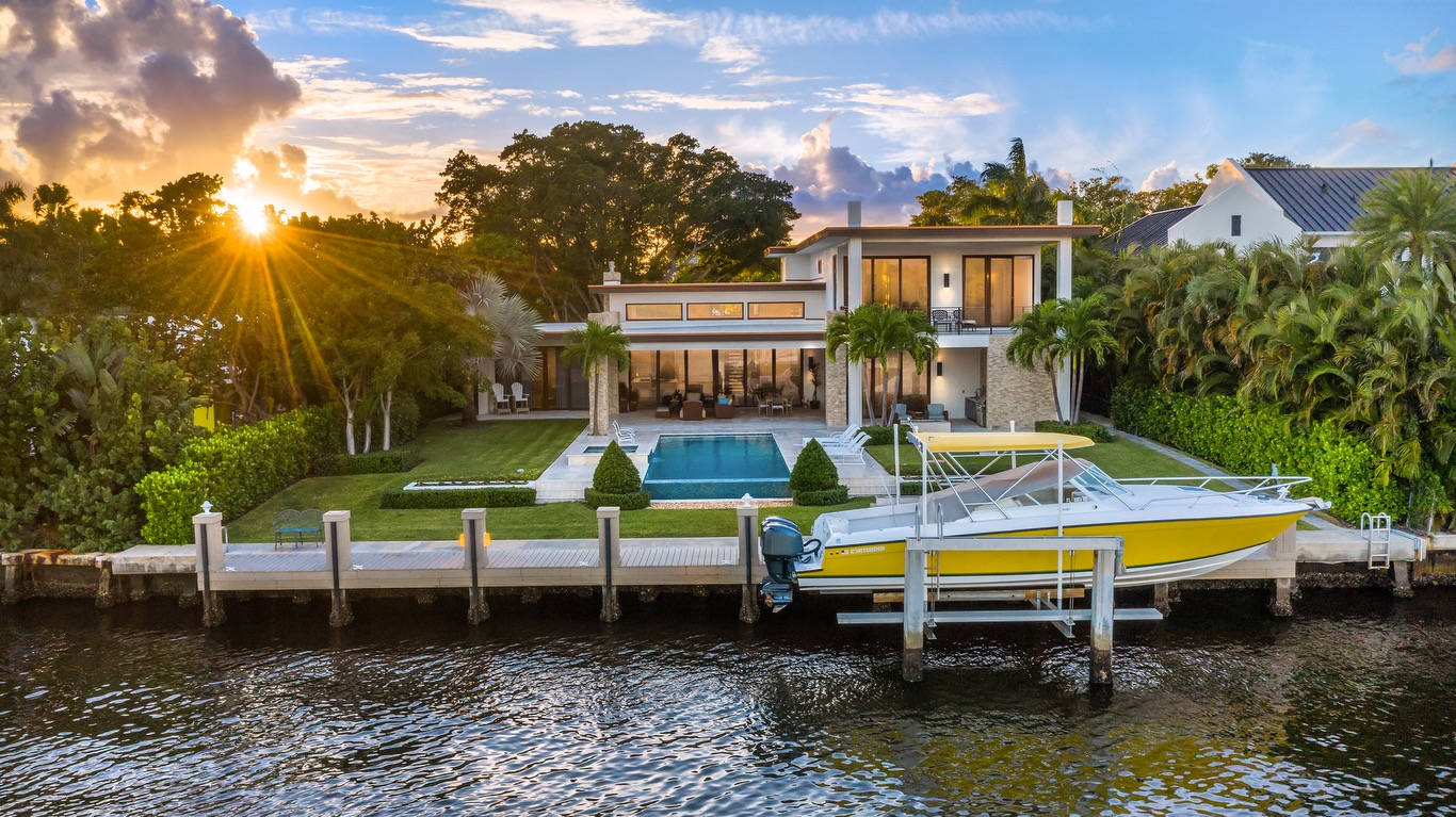 Luxury waterfront house with pool, boat dock, and palm trees during sunset.