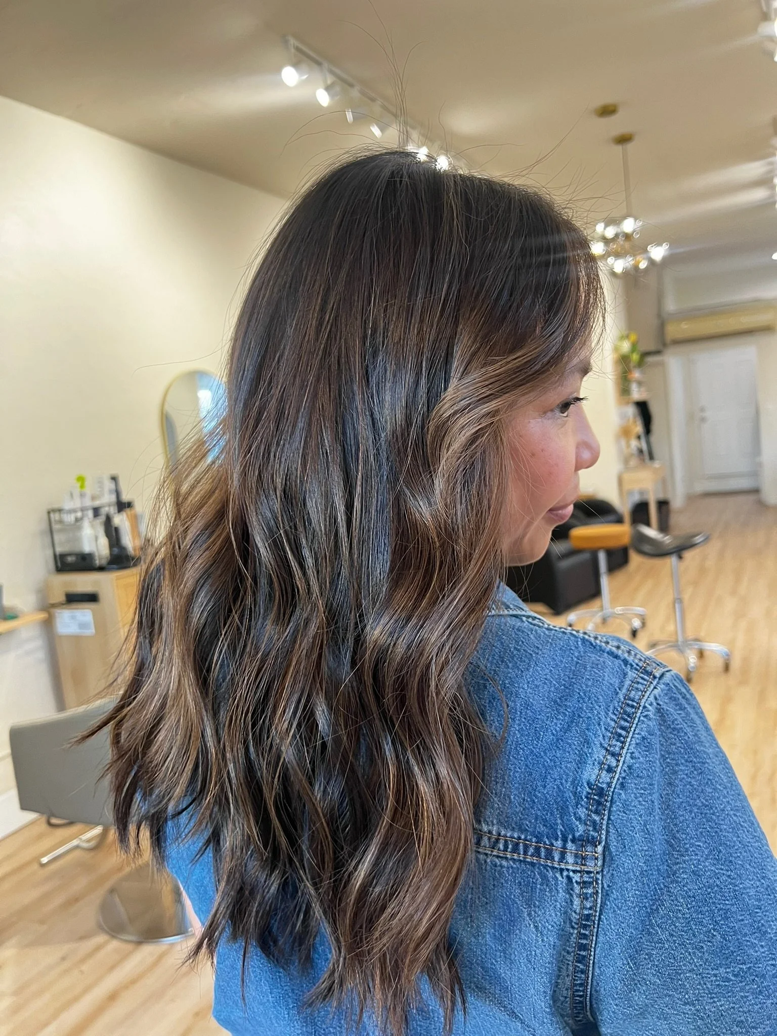 Woman with wavy brown hair sitting in a salon with a mirror, chairs, and hairdressing equipment in the background.