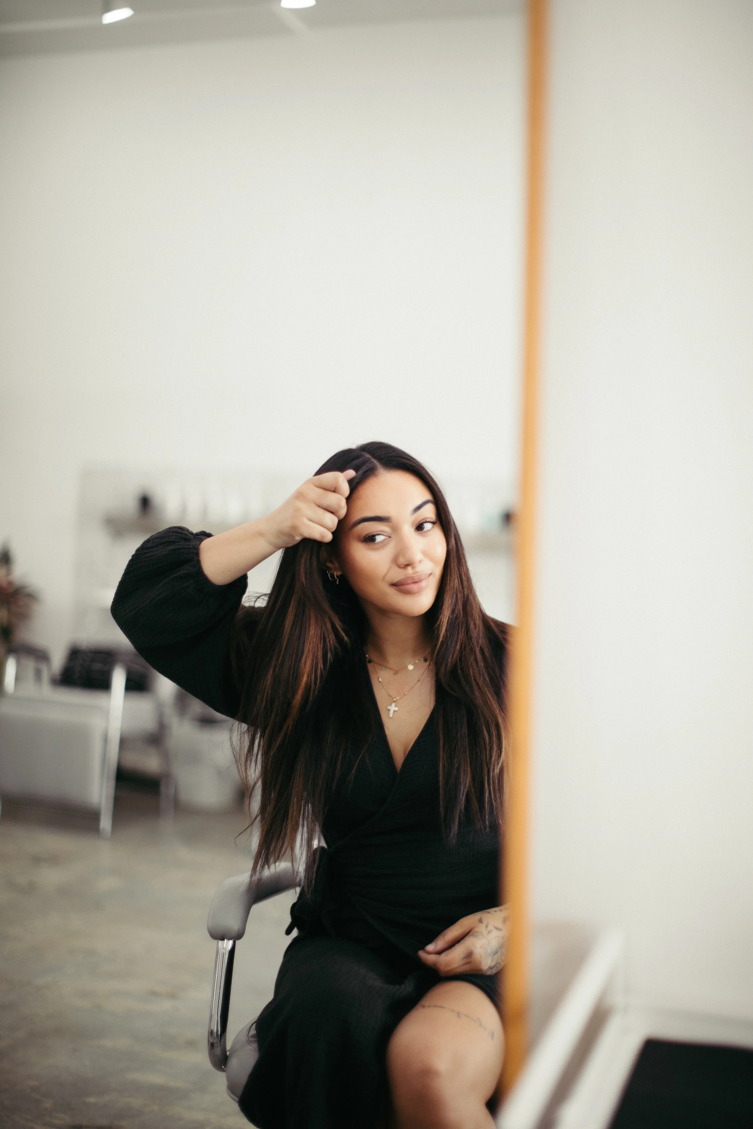 A woman with long dark hair wearing a black dress, sitting in a salon chair, looking into a mirror and touching her hair.