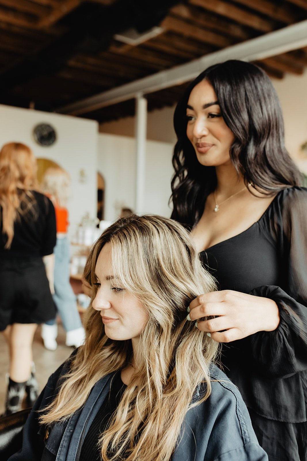 A hairstylist with long black hair styling a woman's wavy blonde hair in a salon with other clients and stylists in the background.
