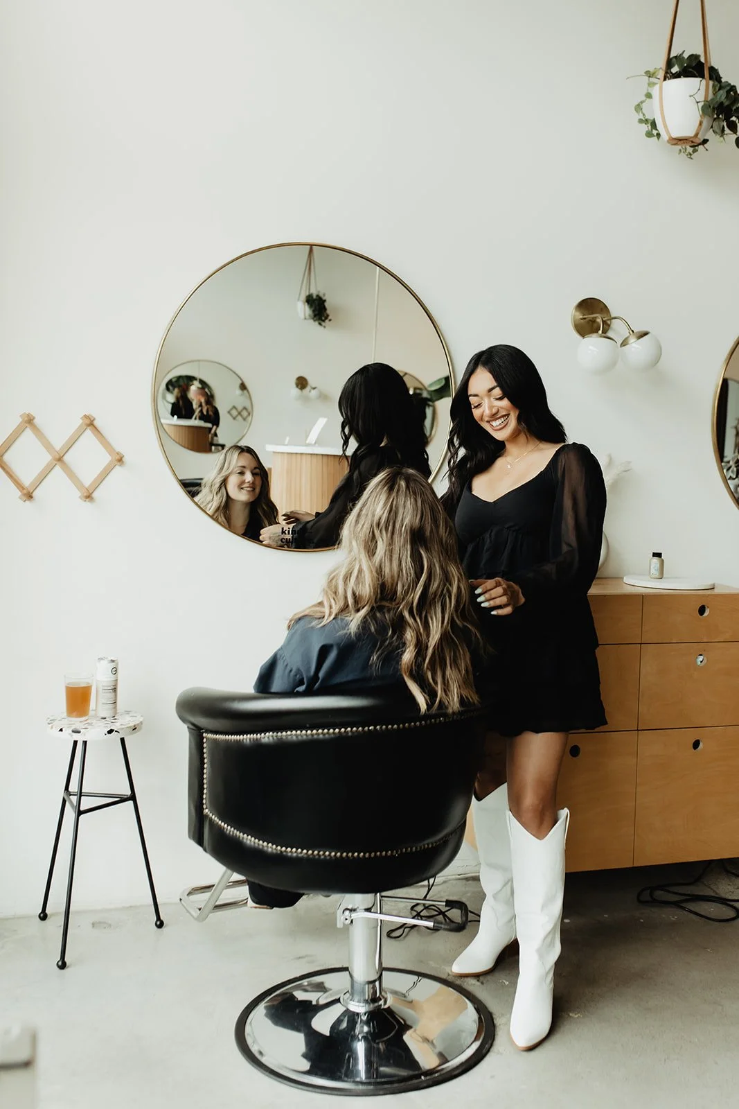 Women in a hair salon, one sitting in a salon chair and two women standing in front of her, smiling. There are mirrors, a side table with a drink, and a wooden cabinet in the background.