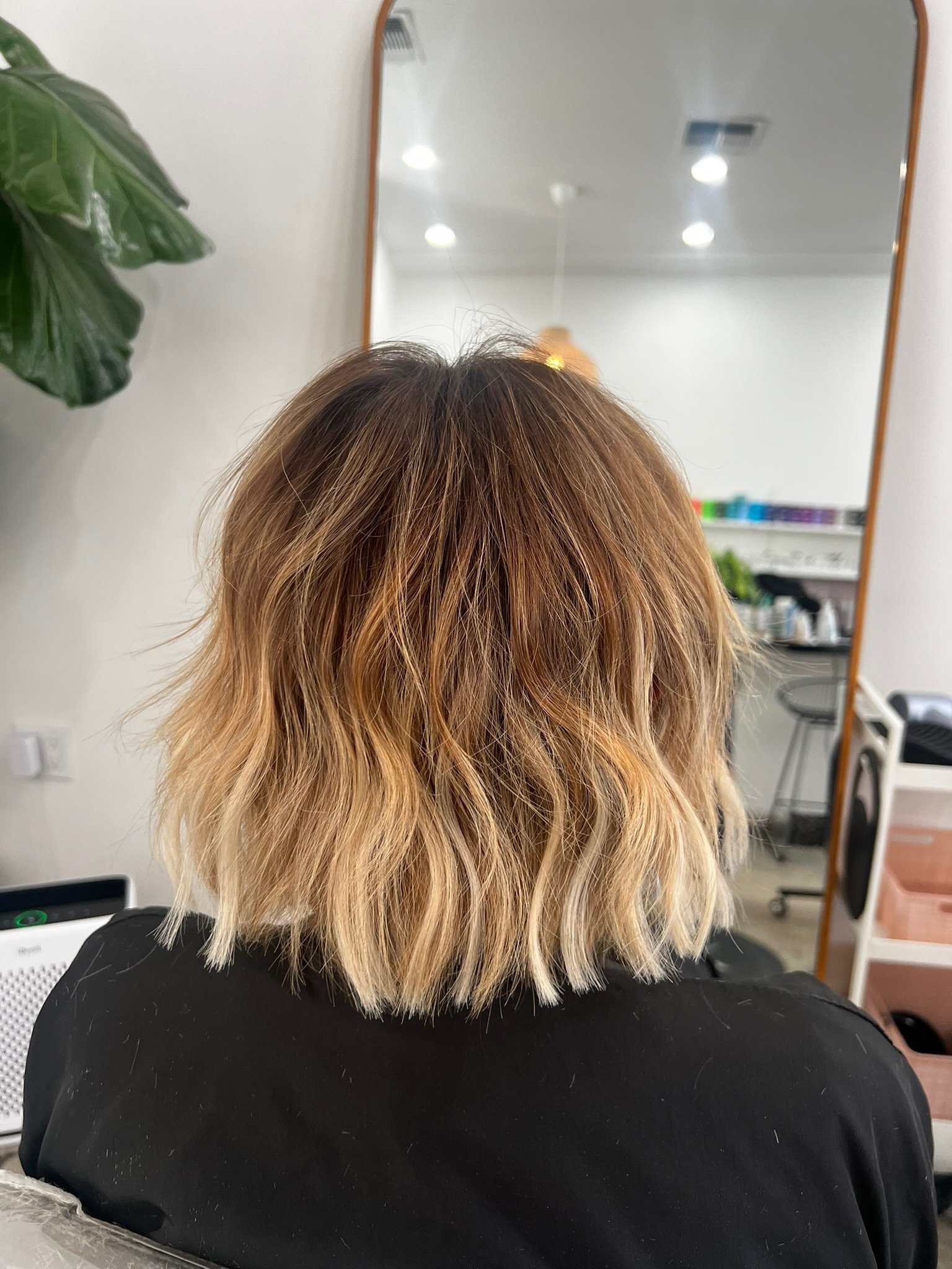 The back of a woman's head with shoulder-length, wavy hair in a balayage style, transitioning from dark brown to blonde, in a salon setting.