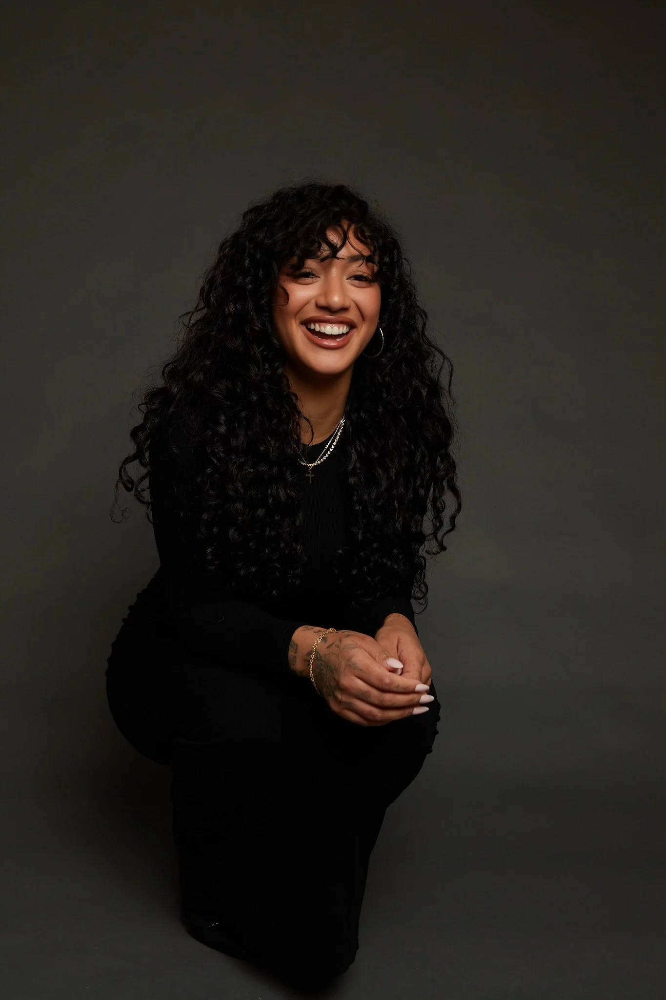 A woman with long, curly black hair, wearing black clothing, smiling, and sitting on her knees against a dark gray background.