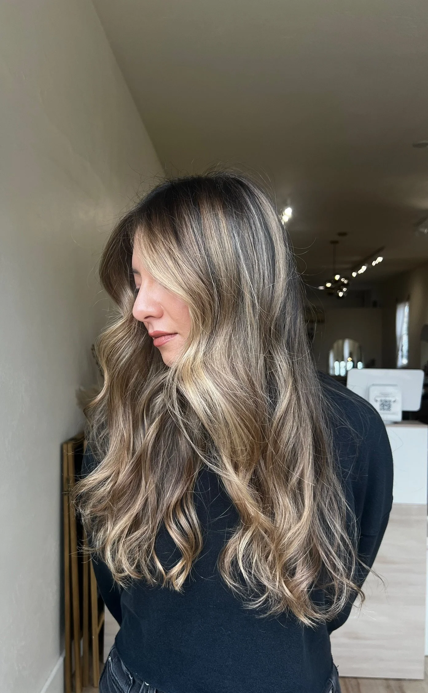 A woman with long, wavy blonde hair and dark roots, wearing a black top, standing indoors with her head tilted down.