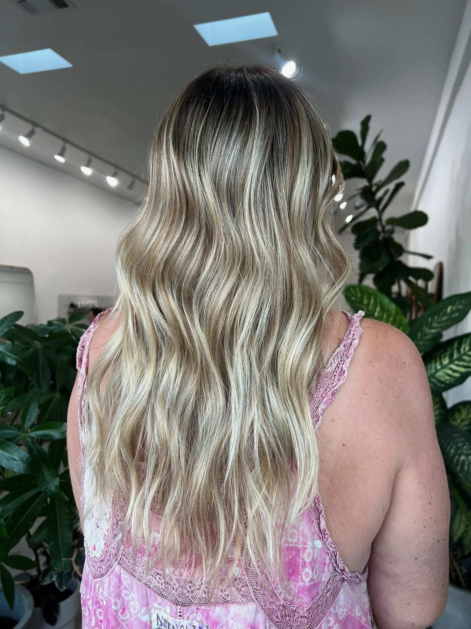 Back view of a woman with long, wavy blonde hair wearing a pink lace top, standing near green plants in an indoor setting.