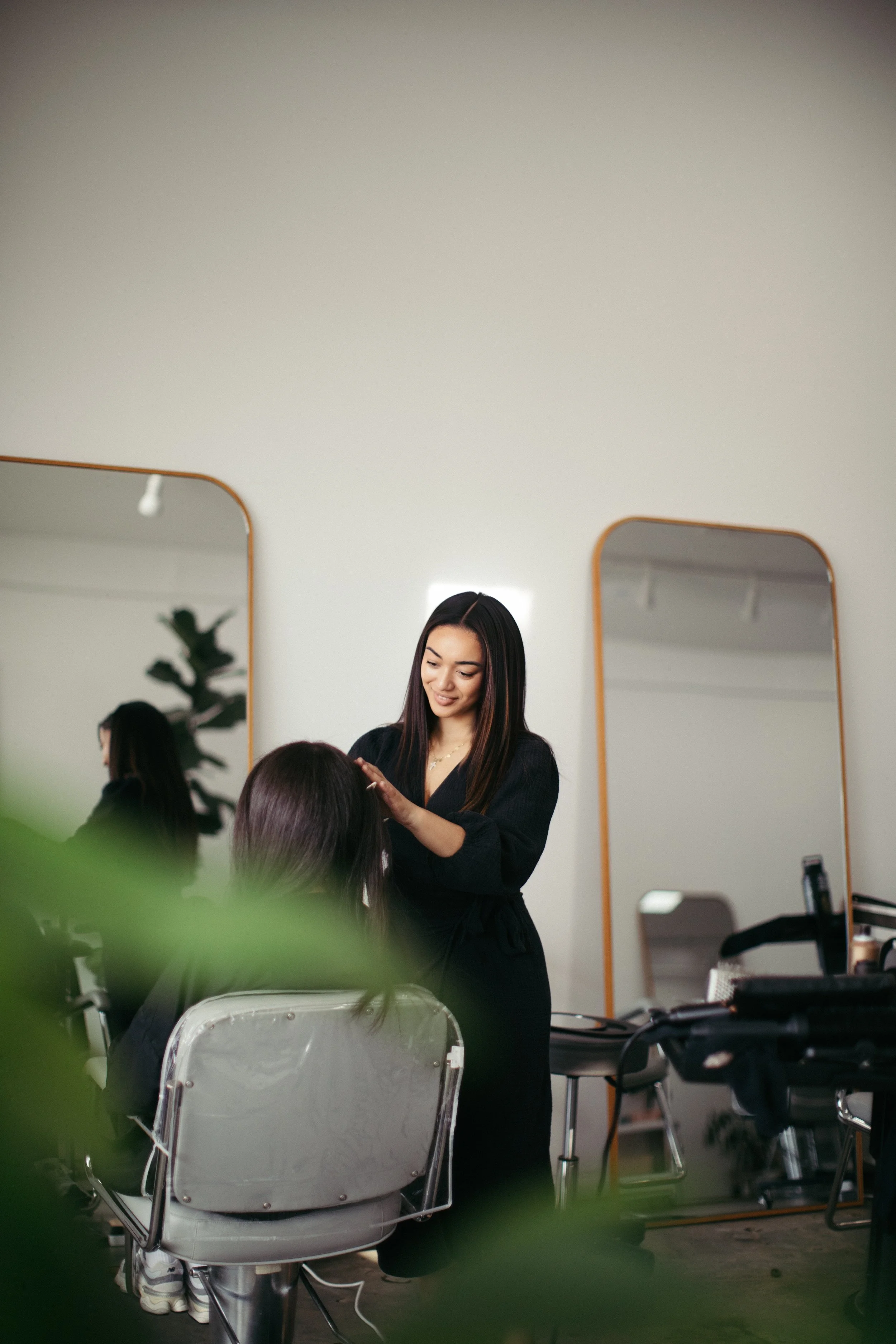 A woman with long dark hair getting her hair styled at a salon, smiling, while a stylist works on her hair in front of two large mirrors.