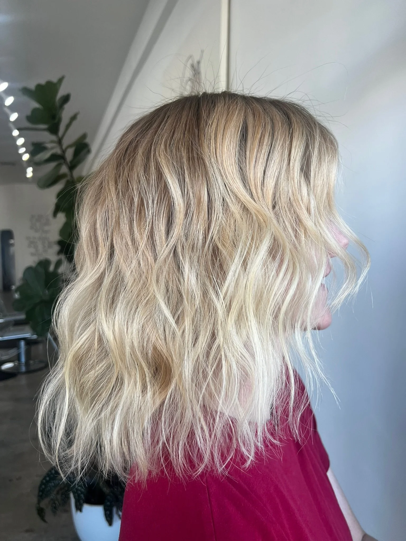 Side view of a woman with blonde wavy shoulder-length hair, wearing a red top, indoors near a plant and white wall.