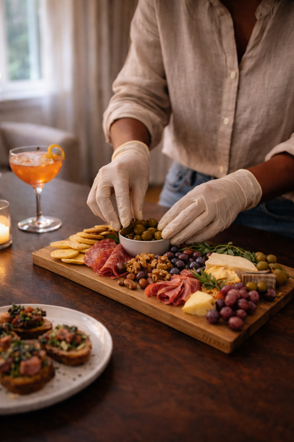 Person preparing a charcuterie board with cheese, meats, nuts, grapes, and olives, wearing gloves, with a cocktail and candles on the table.