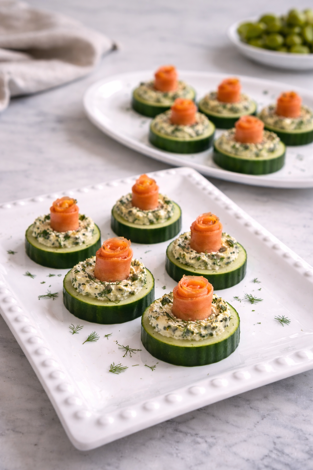 Cucumber appetizer rounds topped with cream cheese, herbs, and slices of smoked salmon rolled into rose shapes, arranged on white rectangular and oval plates.