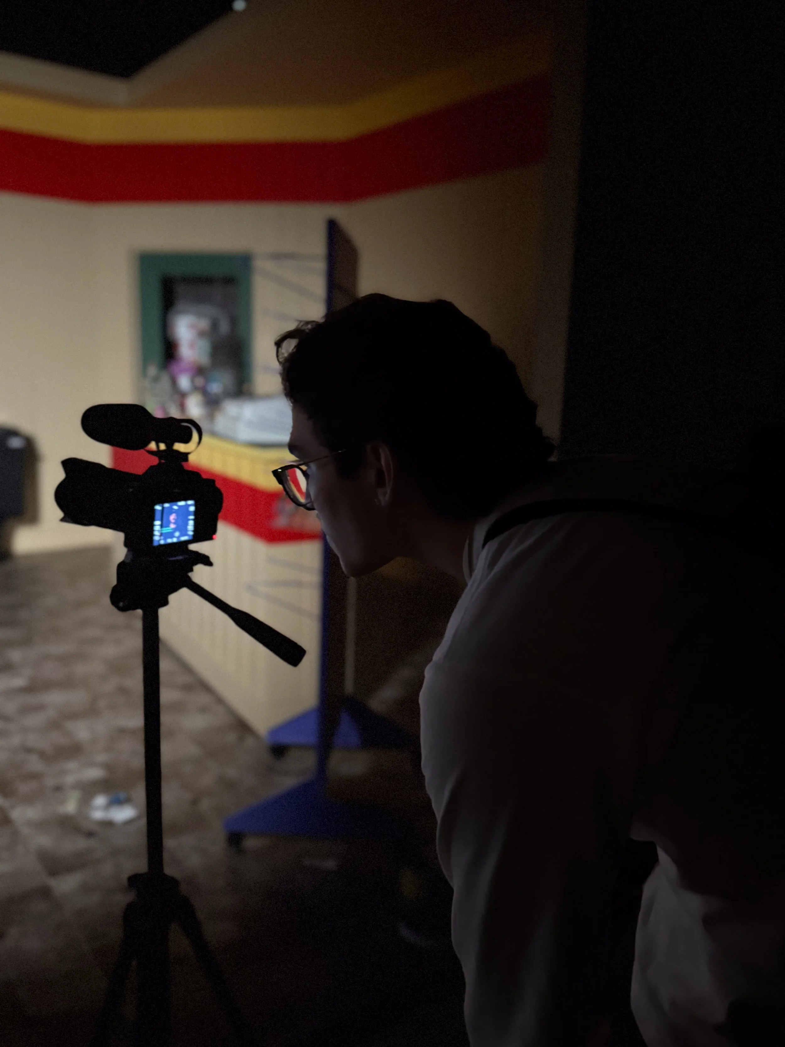 A person with glasses is recording a scene with a camera on a tripod in a dimly lit room.