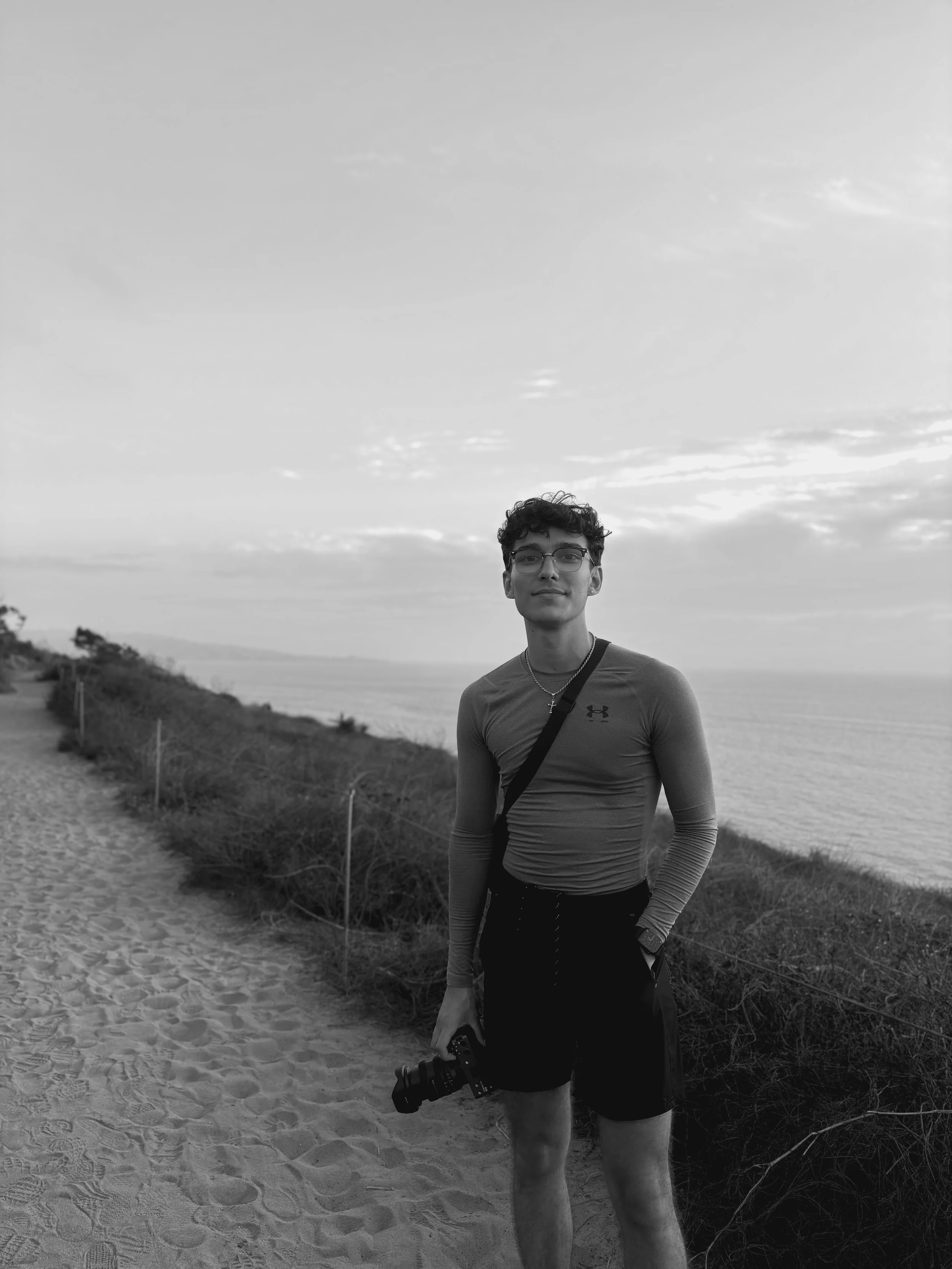 A young man with glasses, curly hair, and a small beard holding a camera while standing on a sandy beach path next to shrubs, with an ocean and cloudy sky in the background.