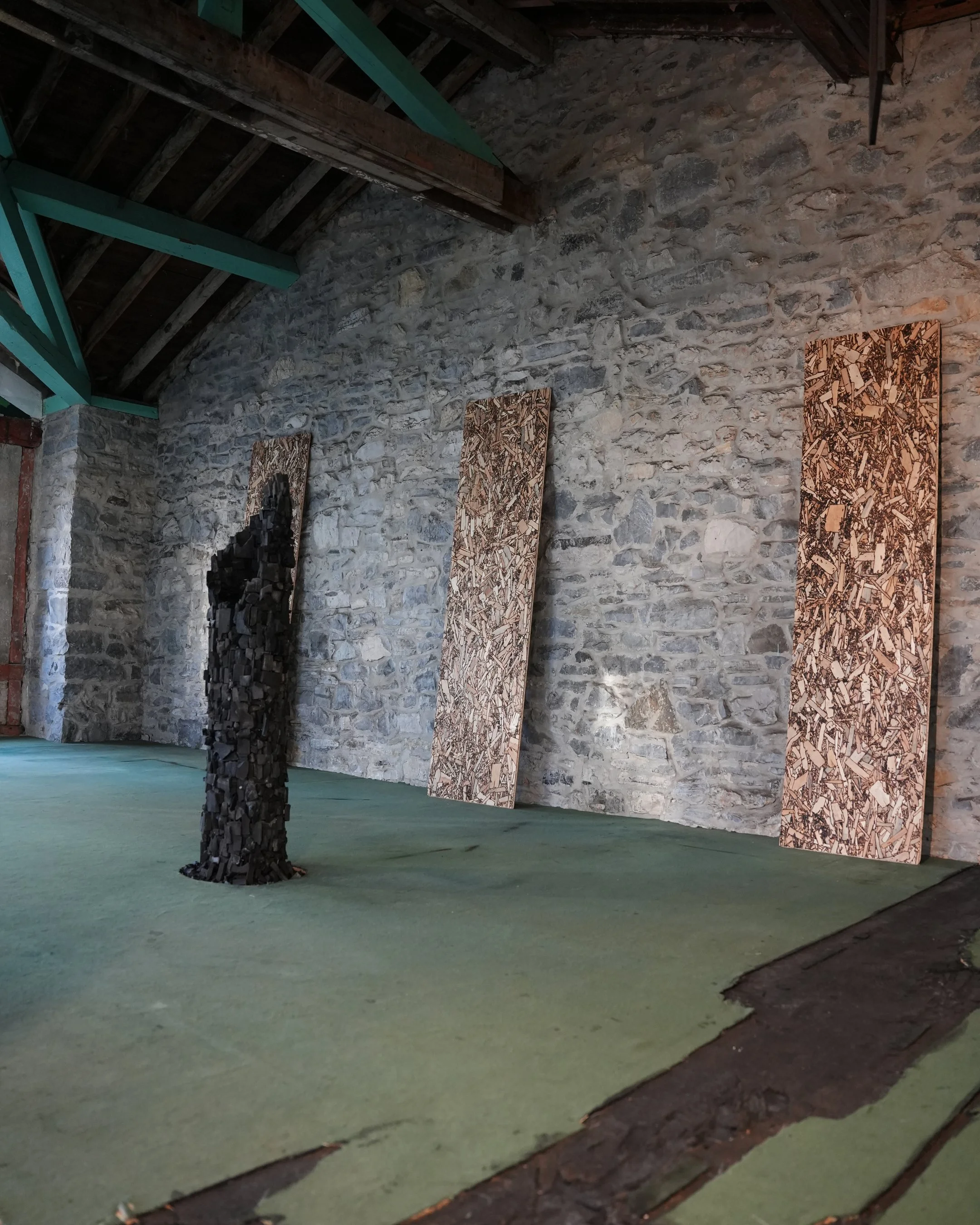 An indoor art installation with a stone wall, three wooden panels with textured patterns, and a black sculpture made of stacked pieces, all on a green floor.