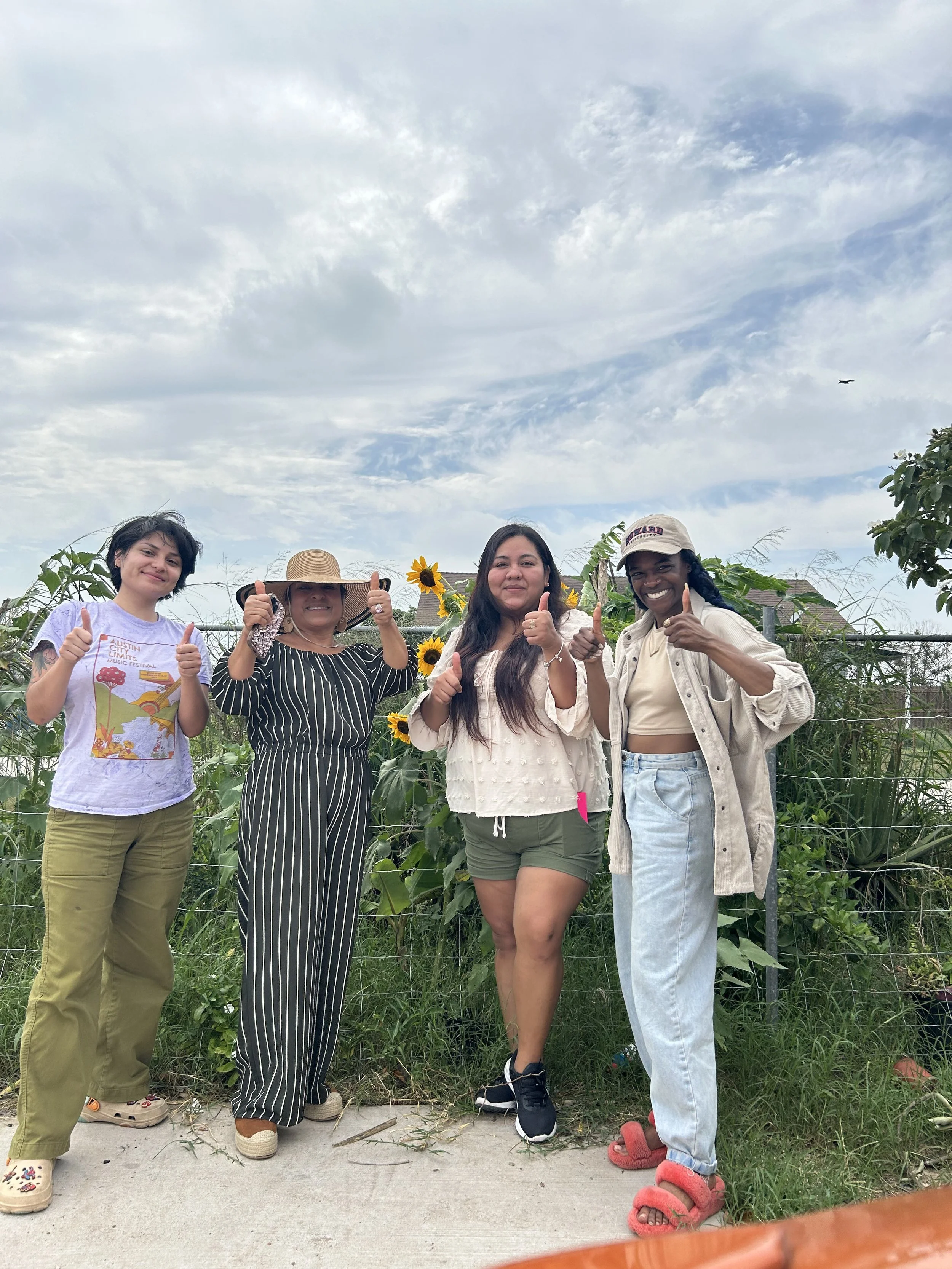 Four women standing outdoors in front of sunflower plants, giving thumbs up and smiling. The sky is partly cloudy.