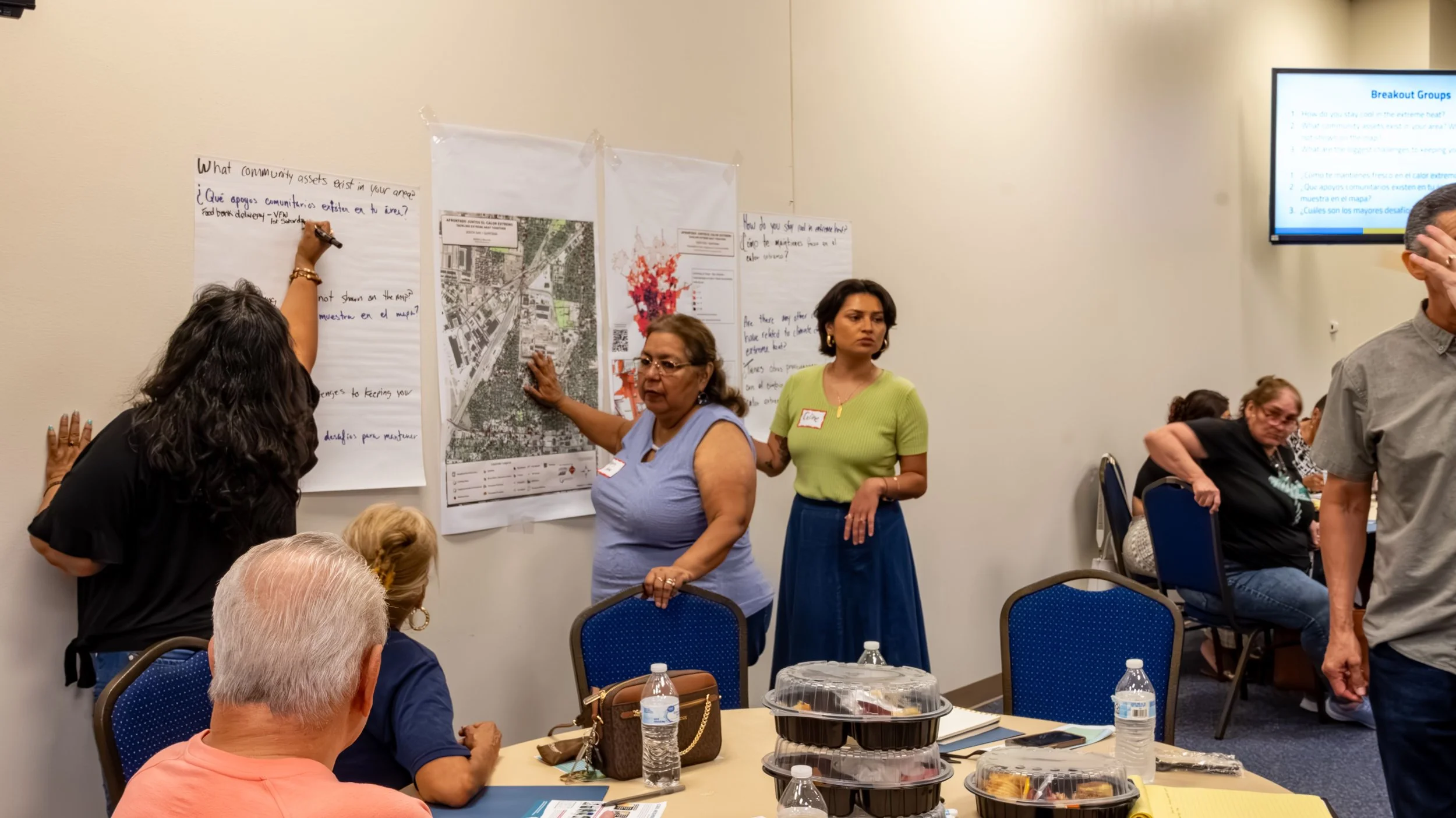 People at a workshop presenting maps and discussion points on a wall with a group seated at tables with food and drinks.