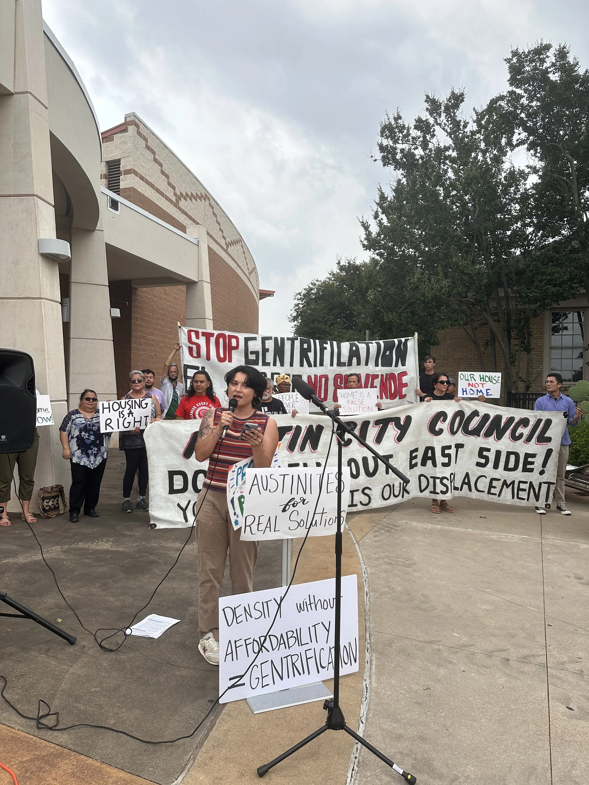 Celine Rendon speaking at a protest rally outdoors, holding a sign that reads 'AUSTIN TEAXAS for REAL Solutions,' with a group of diverse protesters behind her holding banners and signs advocating against gentrification and displacement.