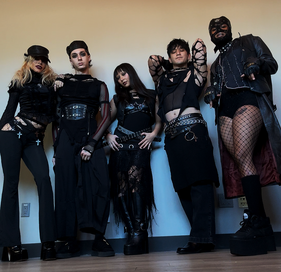 Group of five individuals dressed in punk/goth style clothing, standing against a plain wall, with diverse expressions and poses.