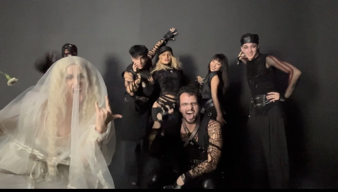 Group of six people dressed in goth or punk fashion, posing against a dark background, one person with a veiled face and someone making a rock gesture.