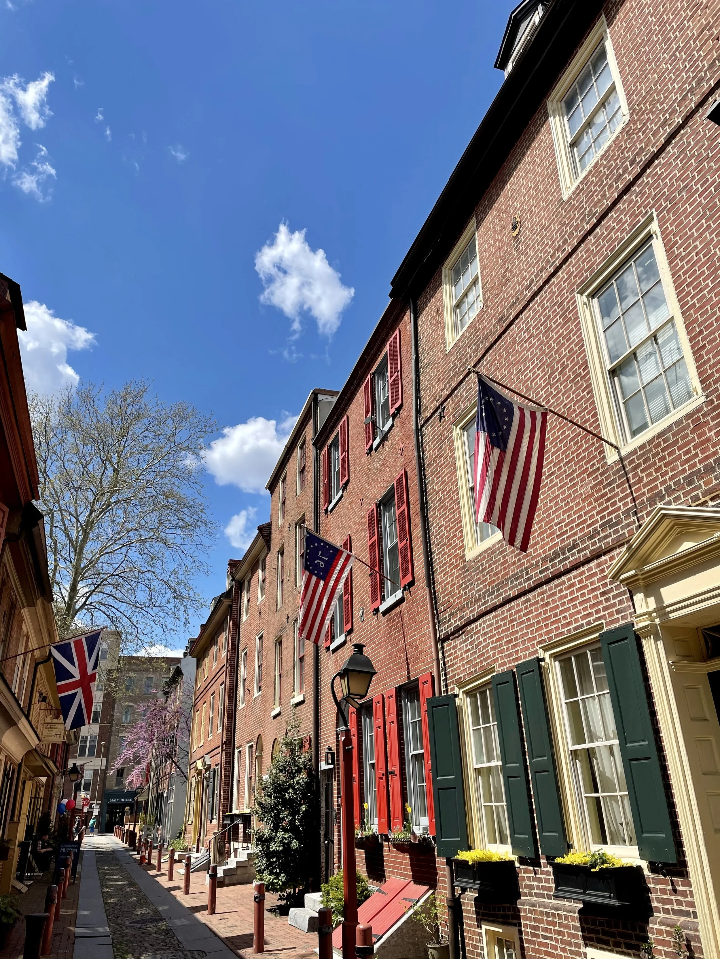 A narrow city street with brick buildings on each side, American flags hanging from poles, green window shutters, a vintage street lamp, blooming pink trees, and a bright blue sky with clouds.
