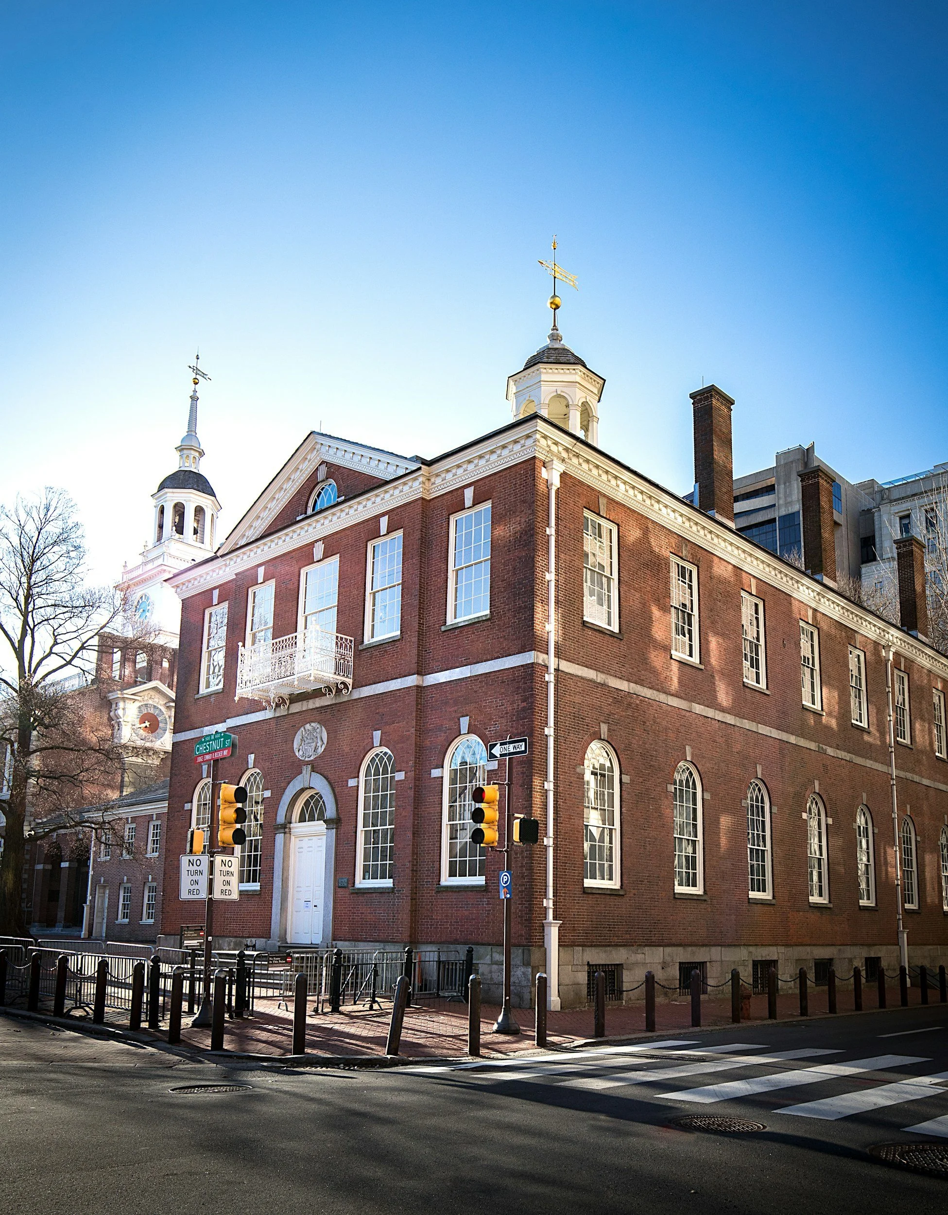 A historic brick building with tall arched windows on a city street corner, featuring a white door, a small balcony, and a clock tower in the background against a clear blue sky.