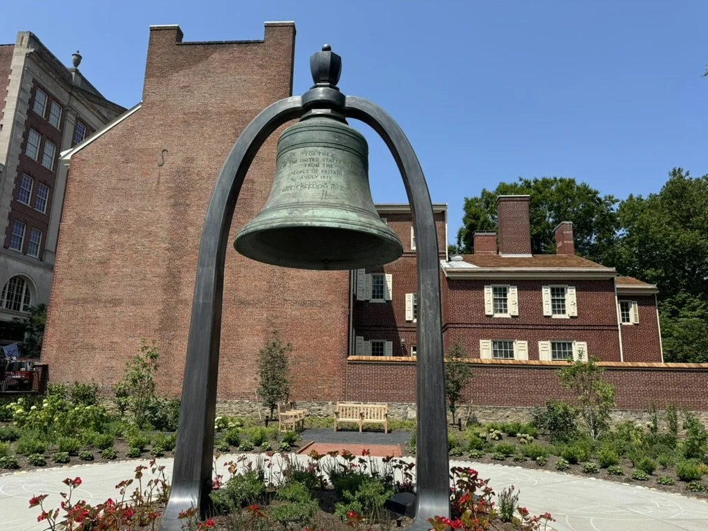 A large bronze bell hanging from a curved metal frame in a small garden, with brick buildings and greenery in the background.