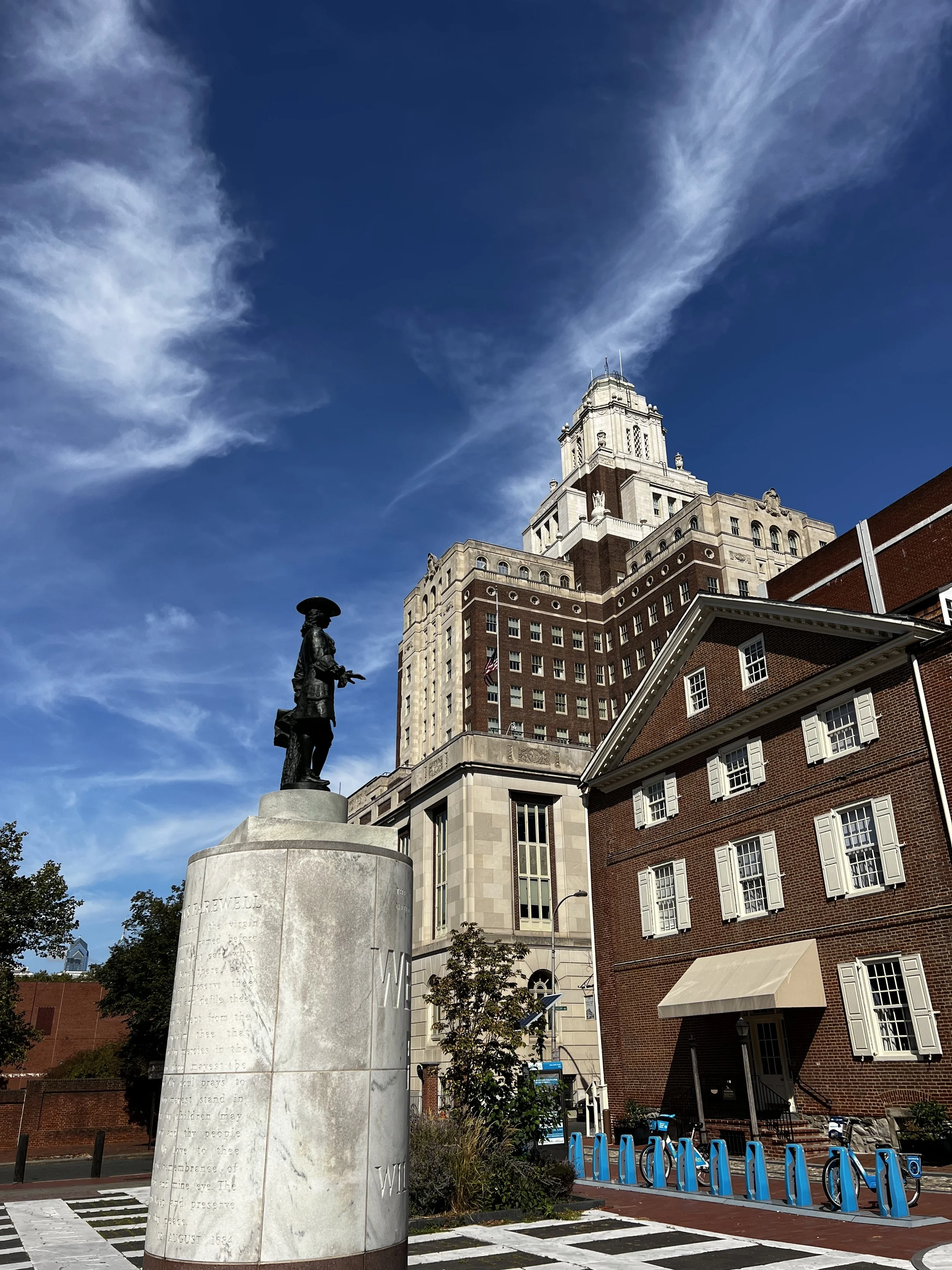 A city scene featuring a statue of a historic figure on a pedestal, a tall building with a spire, a red brick building with white window shutters, and a row of blue bike-sharing stations under a partly cloudy sky.