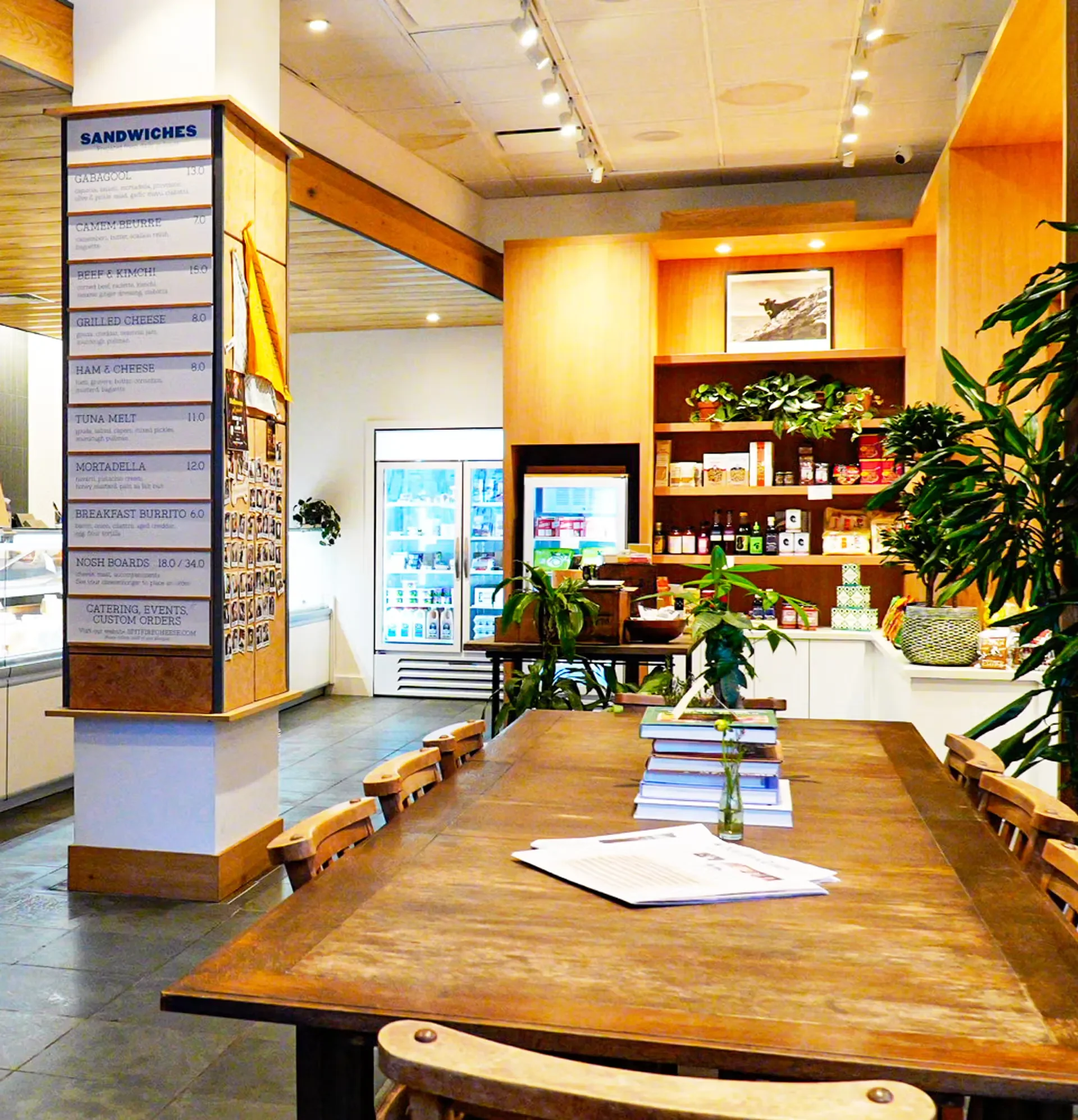 Interior of a cafe or restaurant with a wooden table, chairs, and a shelf displaying food products and books, with a menu board listing sandwich options on the left side.