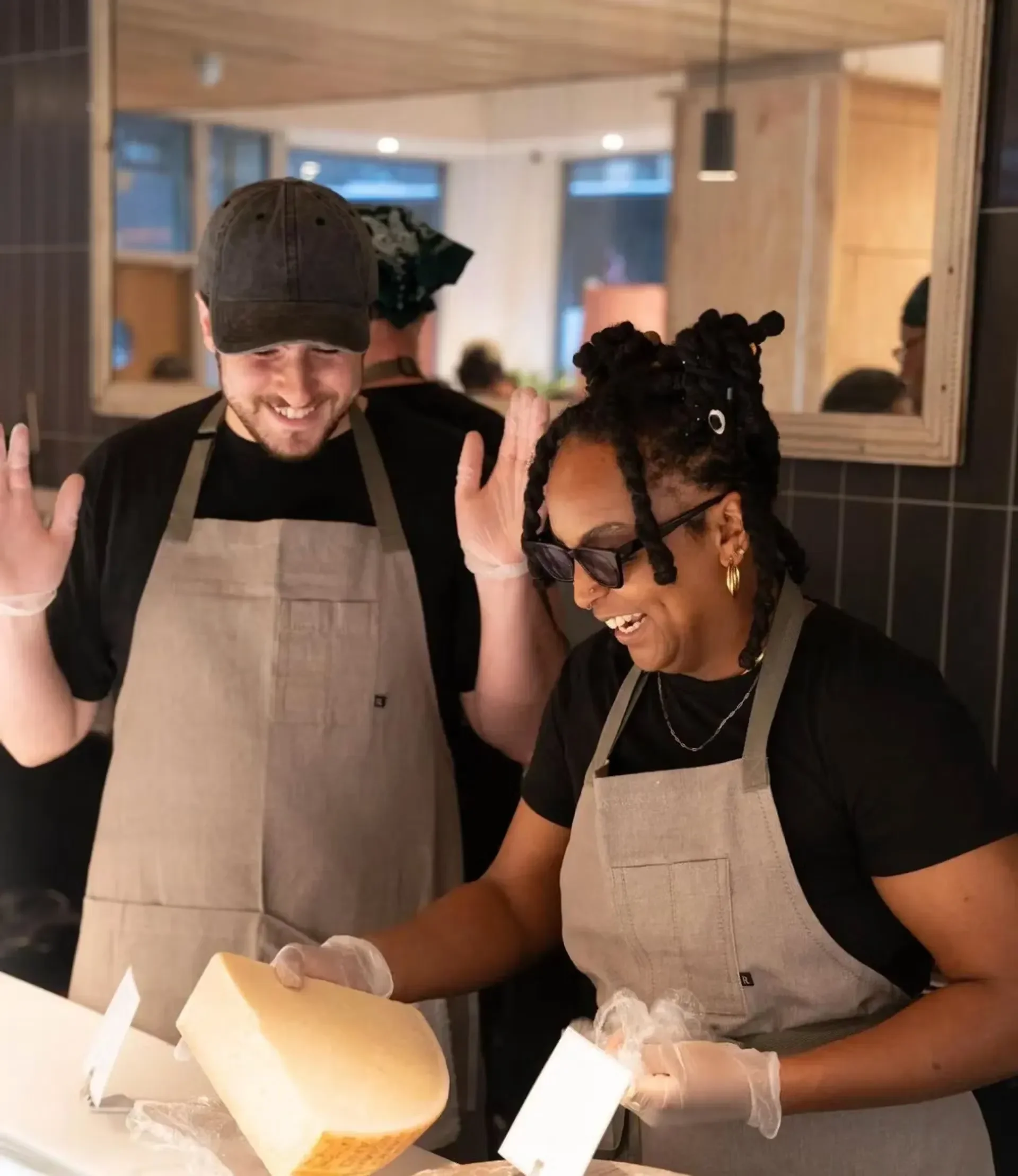 Two people wearing aprons and gloves celebrating with a large block of cheese in a kitchen or restaurant setting.