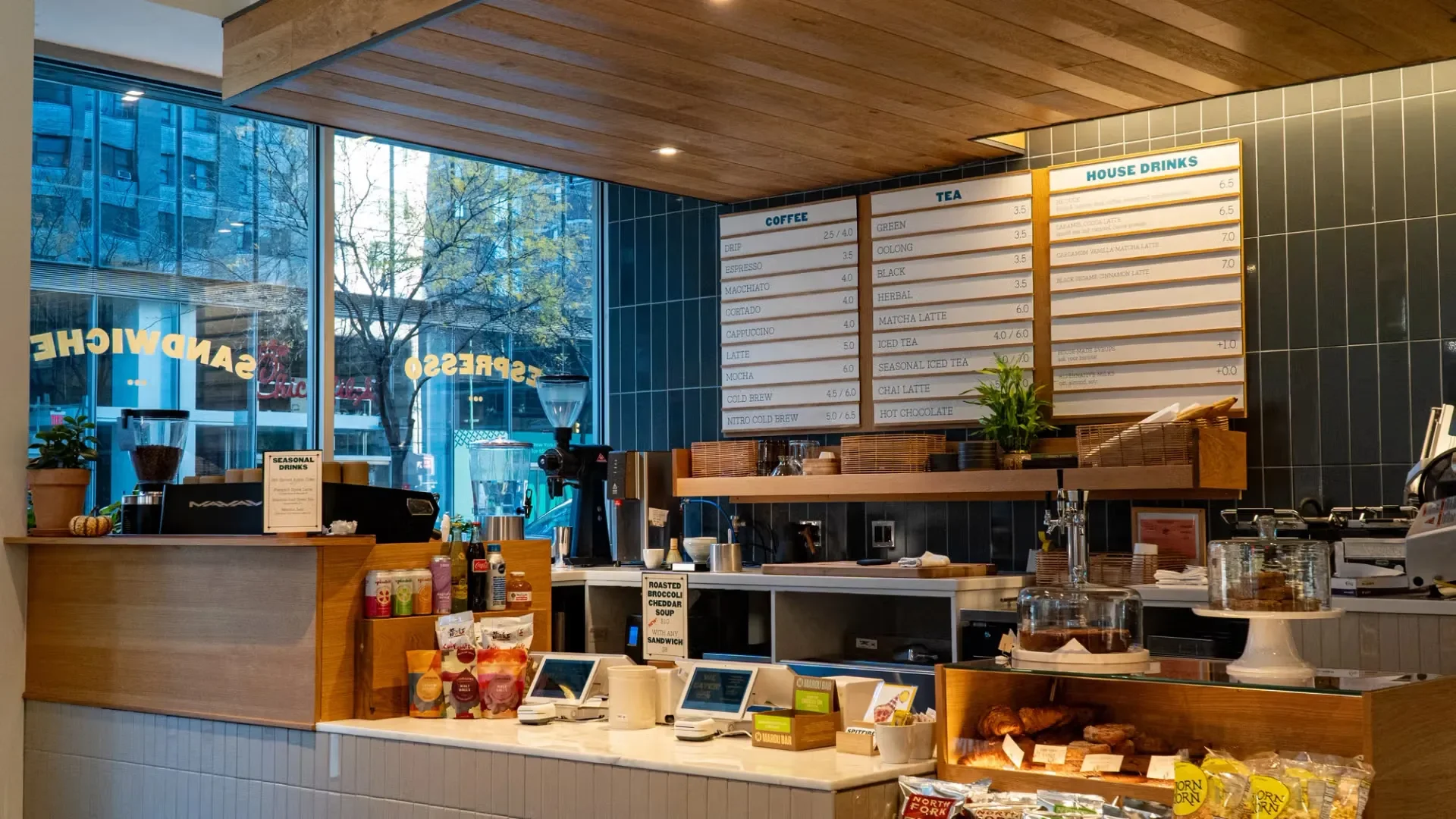 Interior of a coffee shop with a menu board displaying coffee, tea, and house drinks, a wooden counter with condiments, baked goods, and a payment area. Large windows showing trees outside.